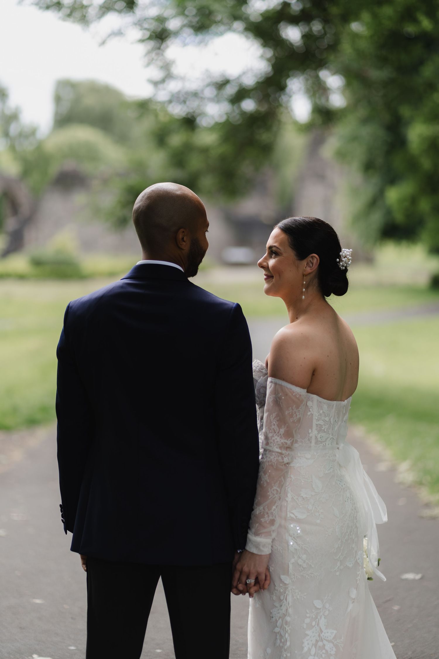 A bride and groom share a candid moment walking together on a tree-lined path during their wedding day.