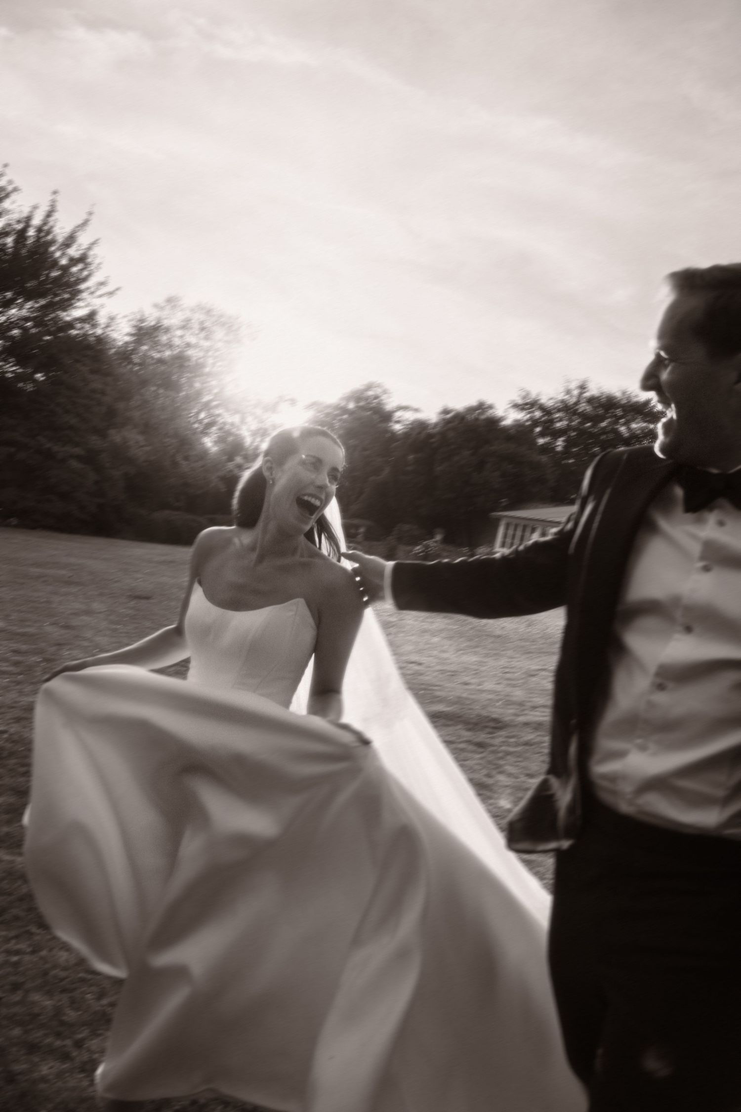 Black and white photo of a joyful bride in flowing gown dancing with groom outdoors on wedding day.
