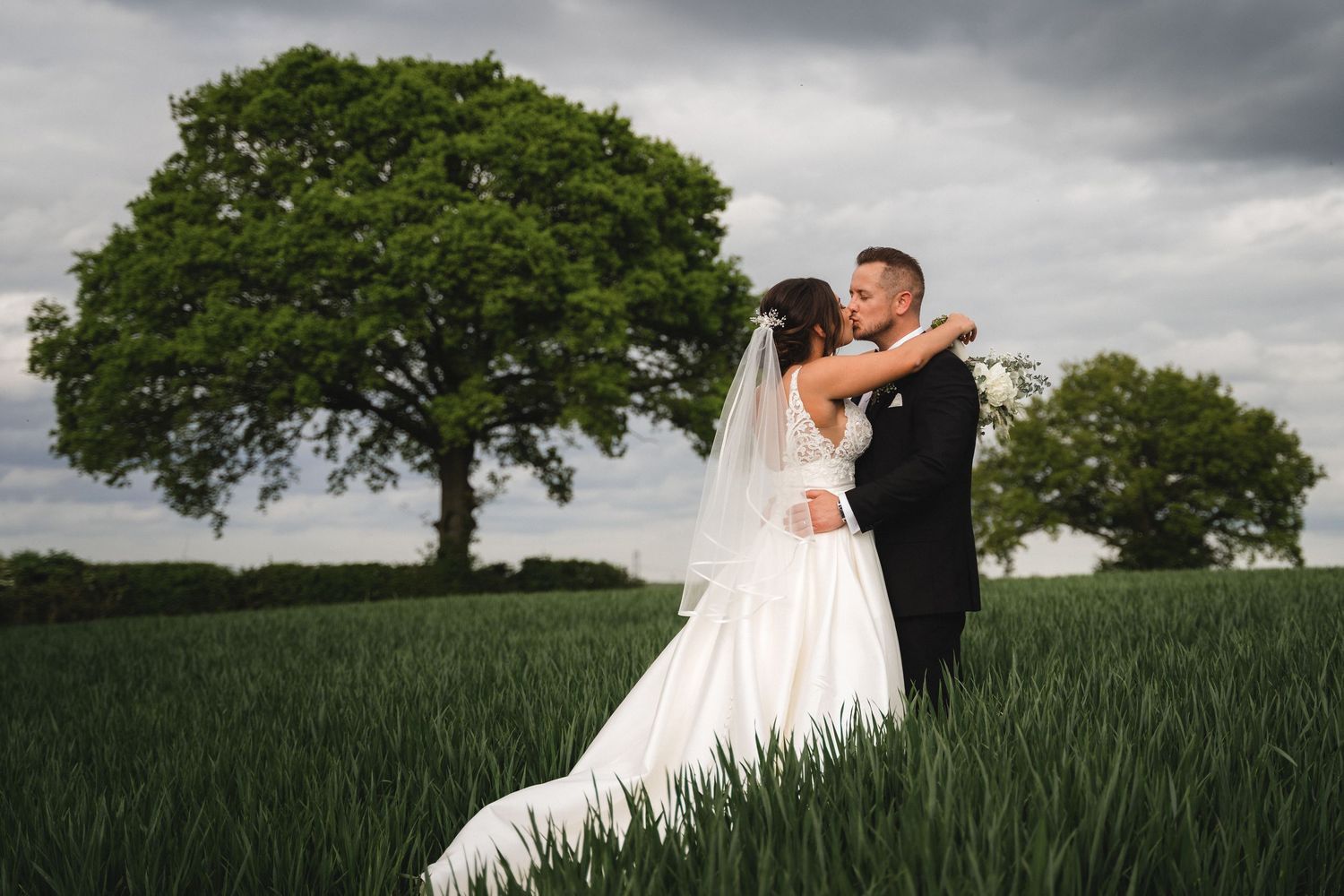 A couple in wedding attire embraces in a lush green field under dramatic storm clouds.