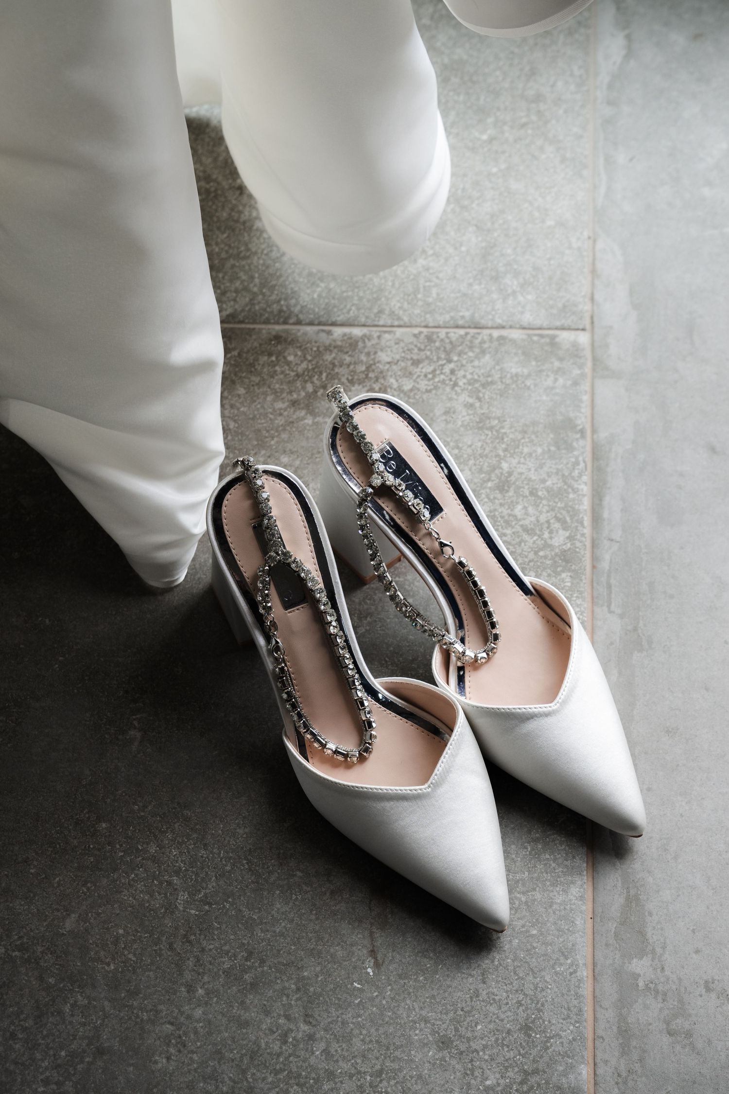 Elegant white pointed-toe bridal slingback heels with crystal embellishments on a gray stone floor.