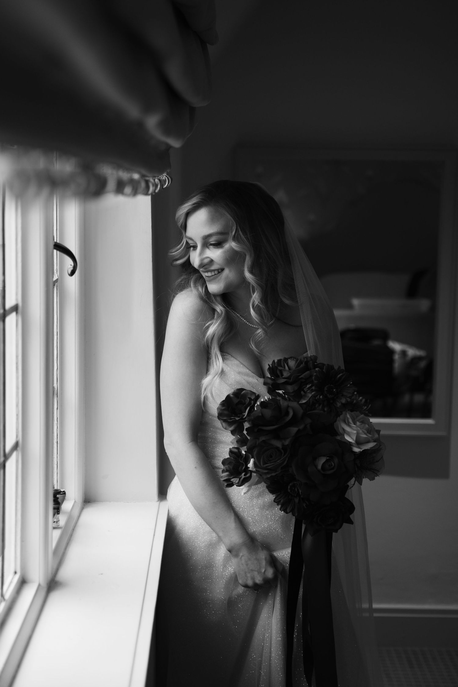 Black and white photo of a bride in a wedding dress standing by a window with dramatic lighting.