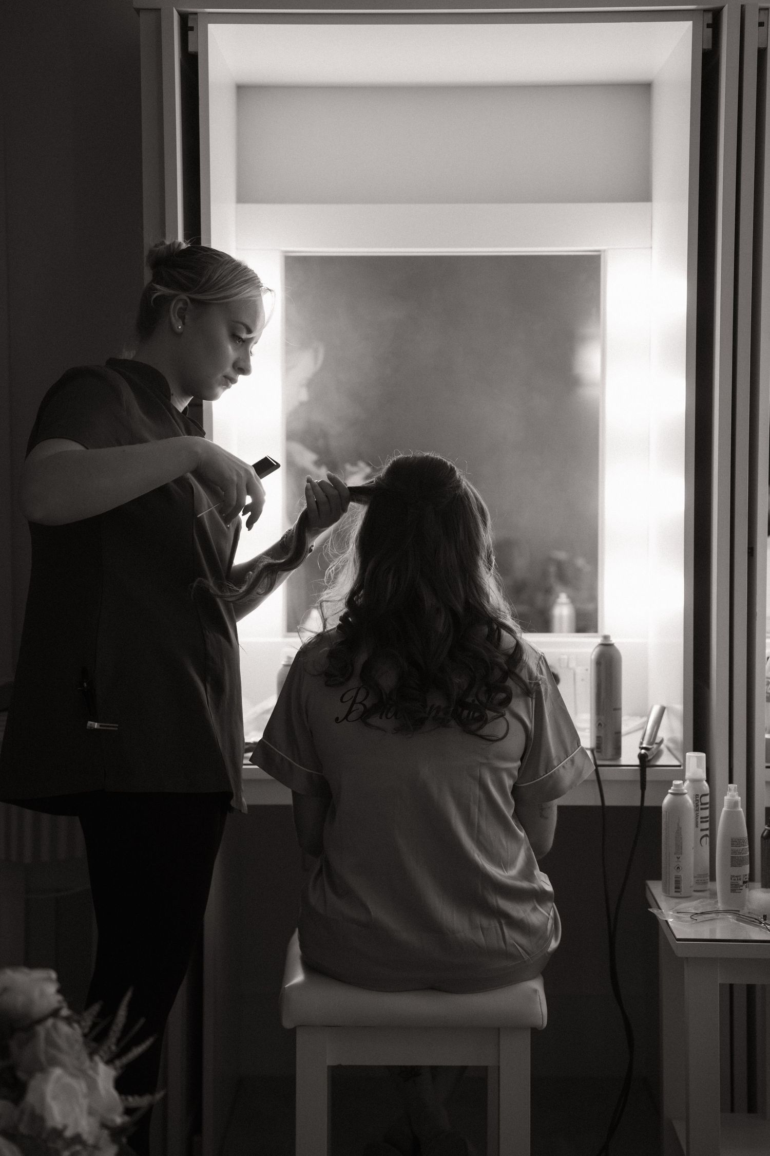 A hairstylist works on styling a client's hair in a black and white photograph with dramatic lighting and mirror reflections.