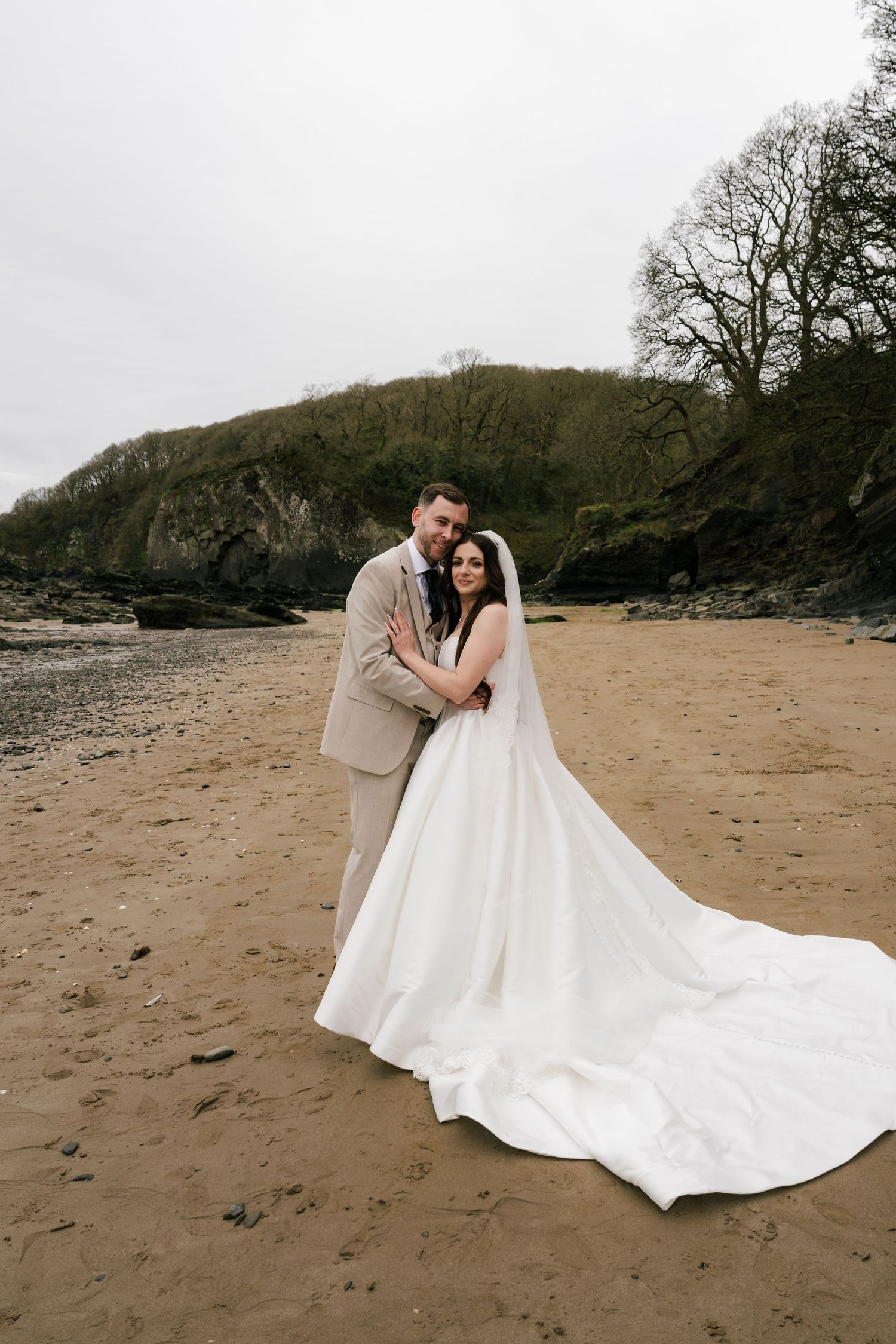 A bride and groom embrace on a sandy beach, she wearing a white gown with long train, he in a beige suit.