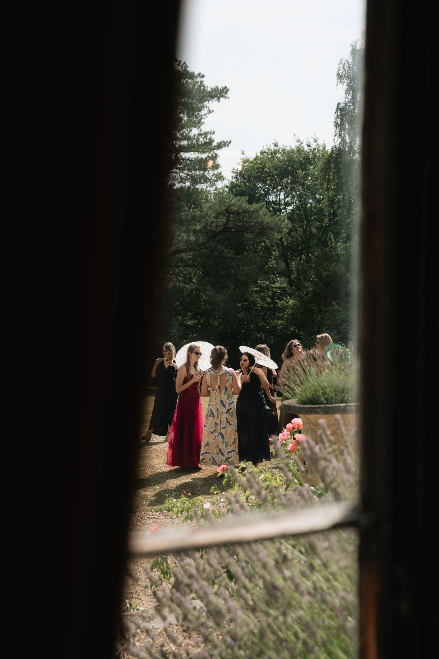 Elegantly dressed wedding guests in colorful gowns and sun hats gather outdoors near lavender blooms on a sunny day.