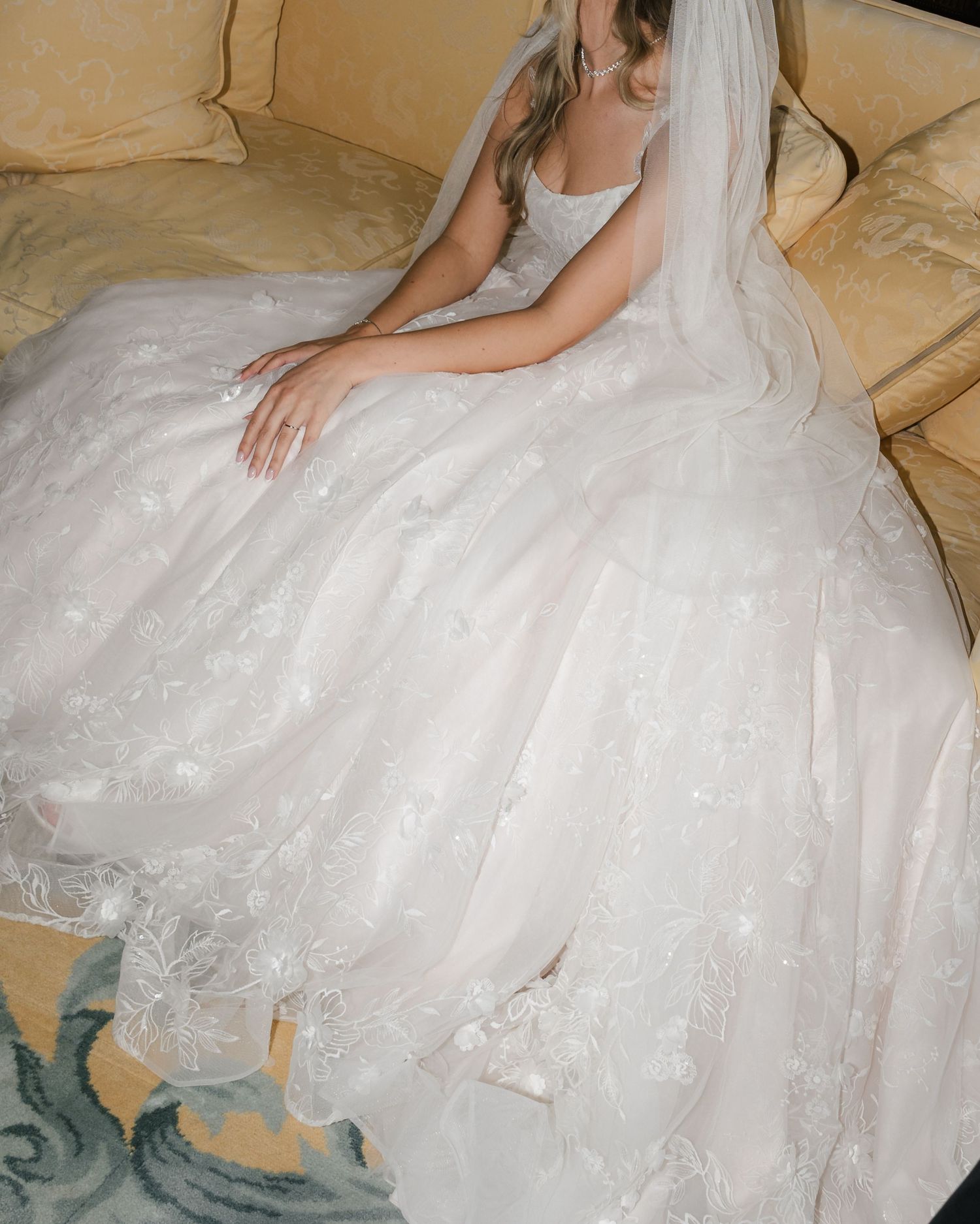 A bride in a flowing white lace wedding gown with tulle skirt sits gracefully on a beige couch.