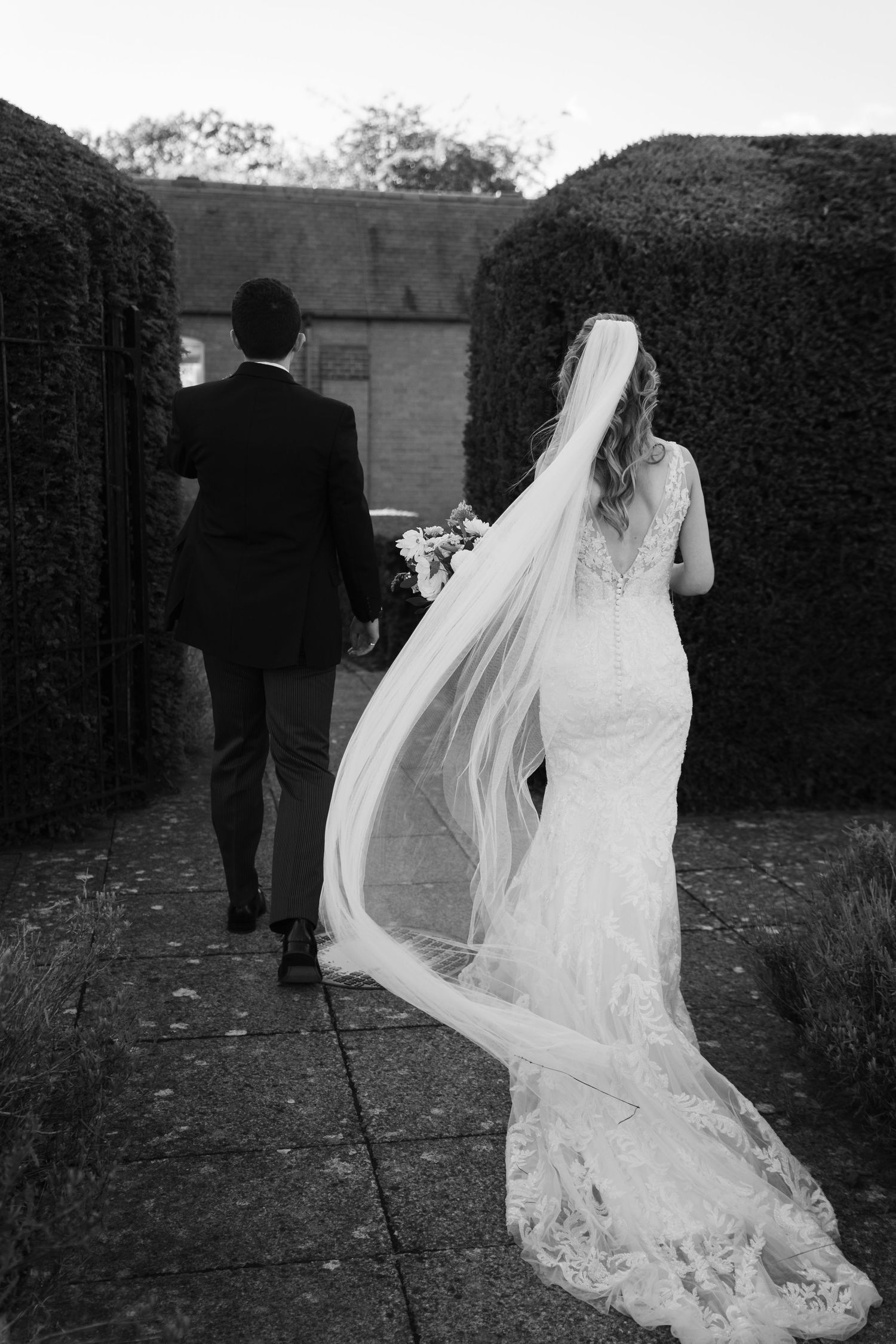 A couple in wedding attire walks through a garden path as the bride's lace veil flows elegantly behind them.