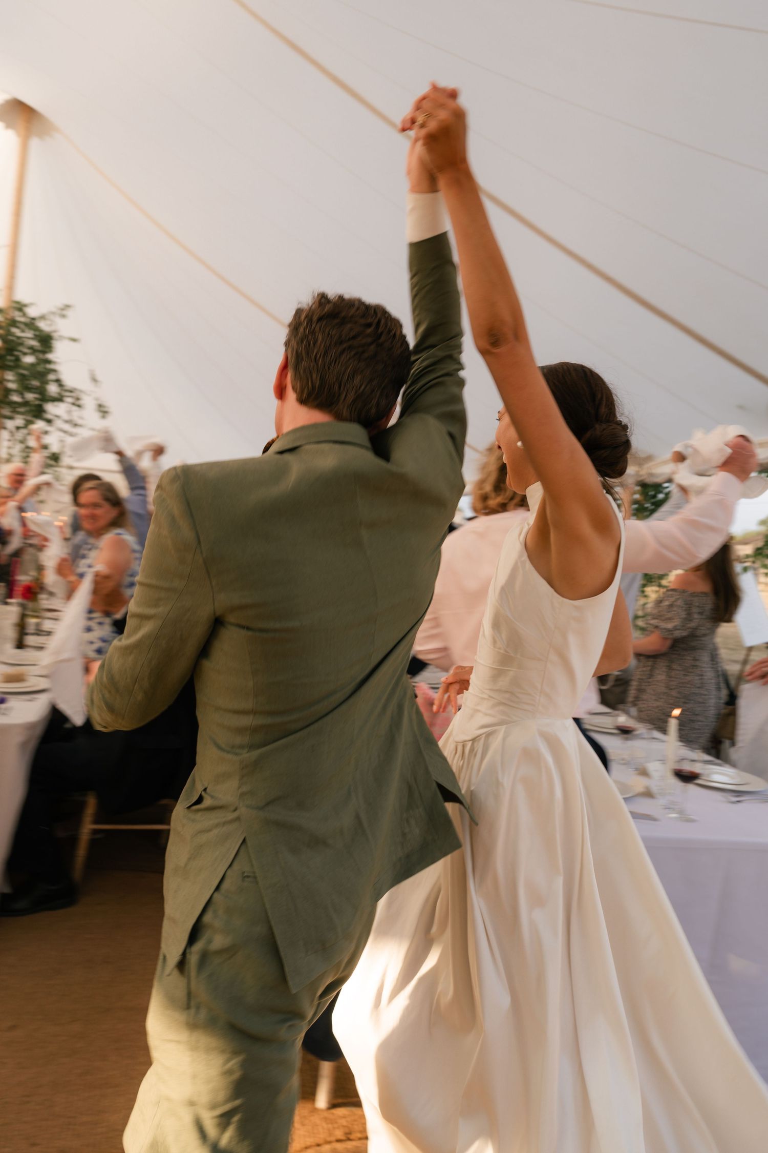 A couple performs their first dance at a wedding reception in a white tent with guests watching in the background.