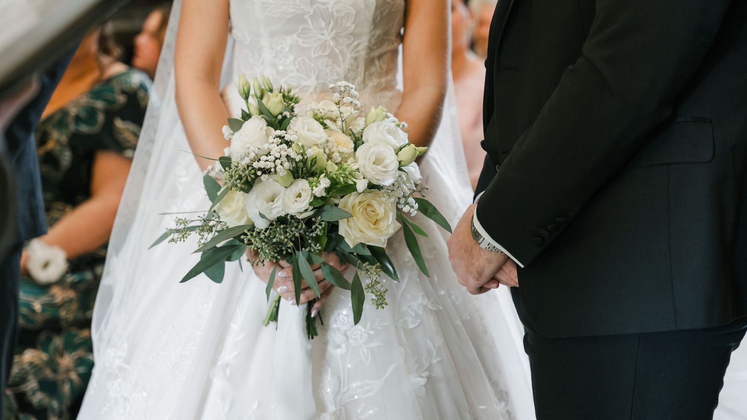 Close-up details of a bride in white wedding dress holding a delicate bouquet with white and green florals.