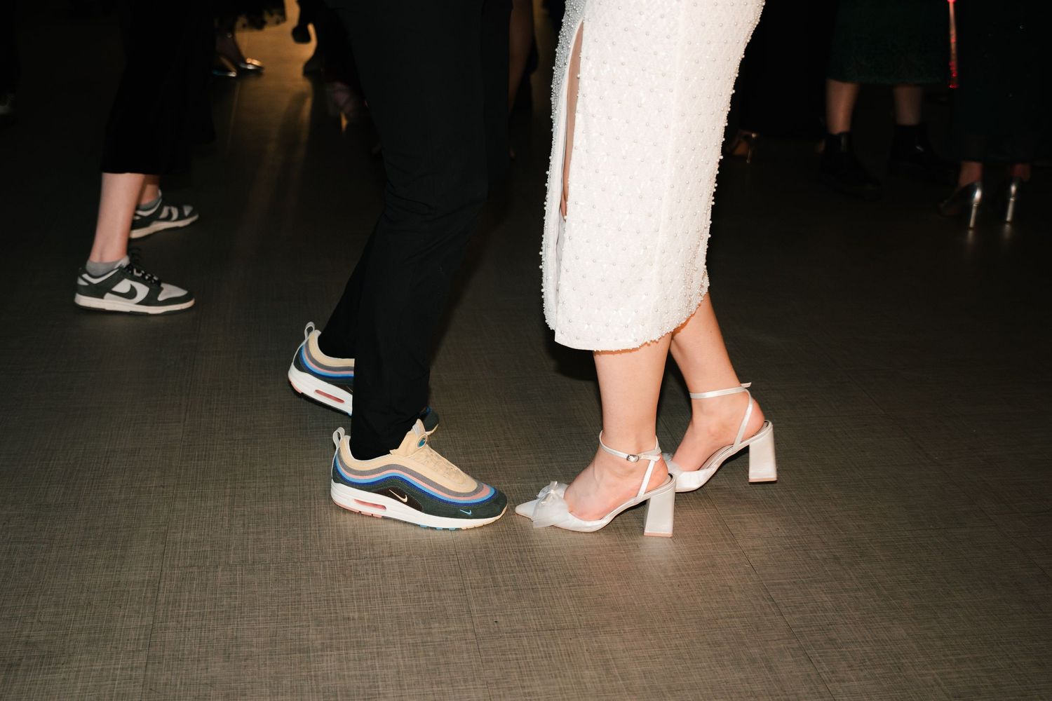 Close-up view of various shoes and feet on a dark floor at a social gathering or event.