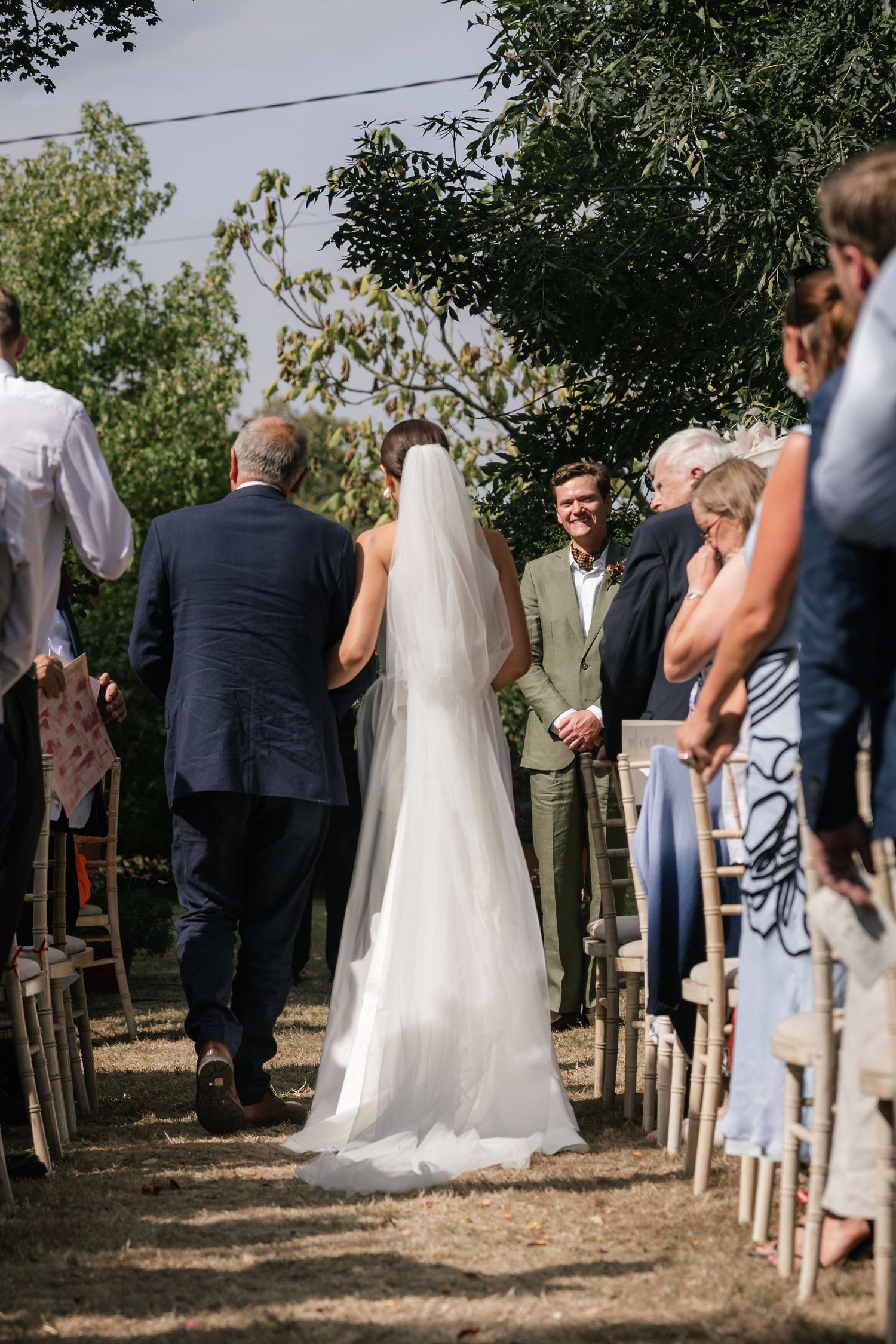 An outdoor wedding ceremony takes place under trees with guests seated on wooden chairs along a dirt path.