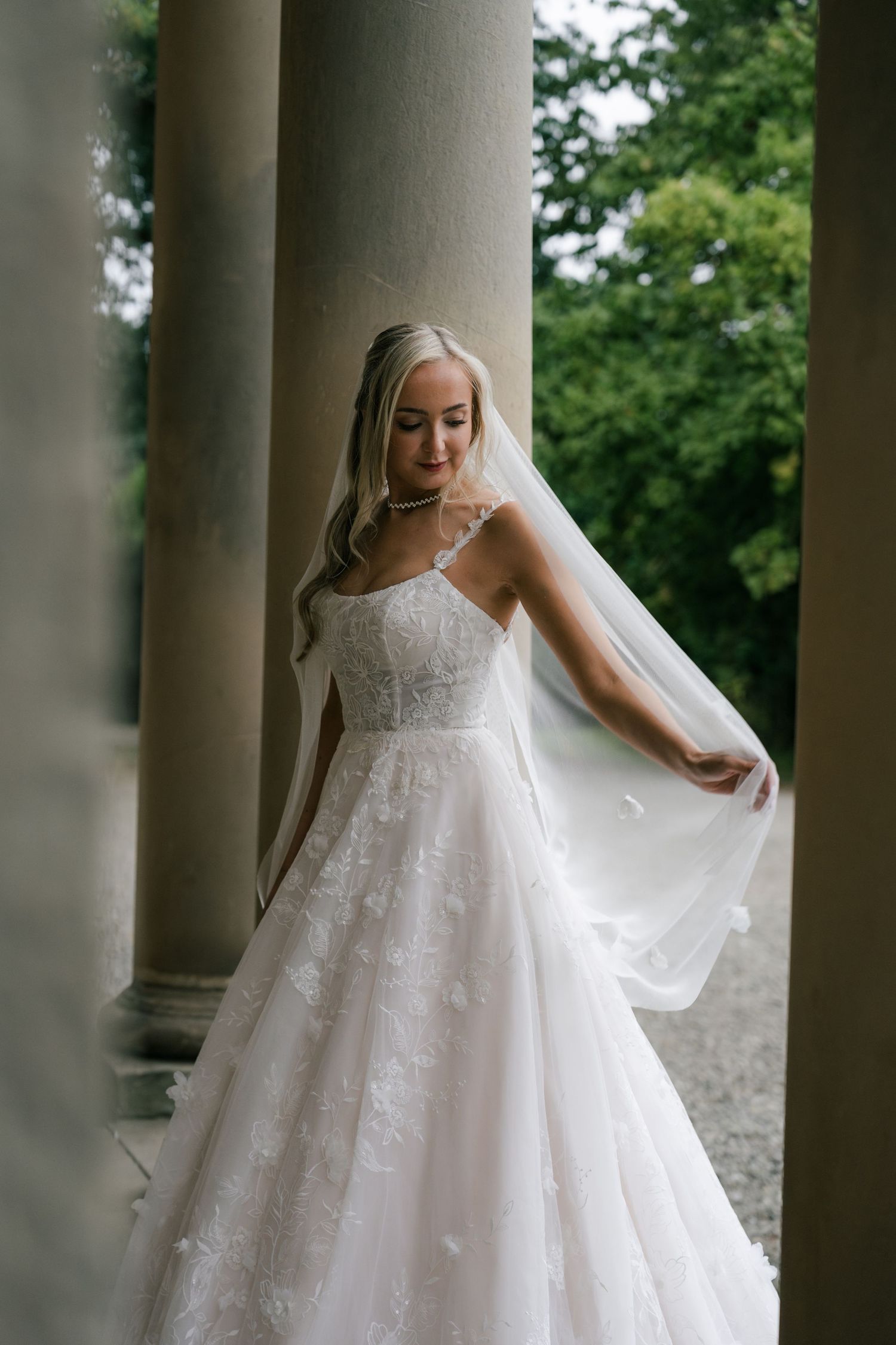 A bride in a flowing white lace gown poses elegantly among classical stone columns.