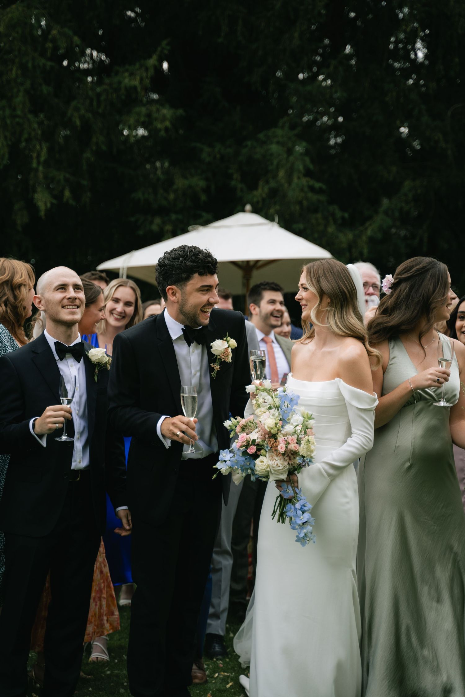 Wedding party members share candid laughs and embraces during outdoor celebration with sage green bridesmaids dresses.