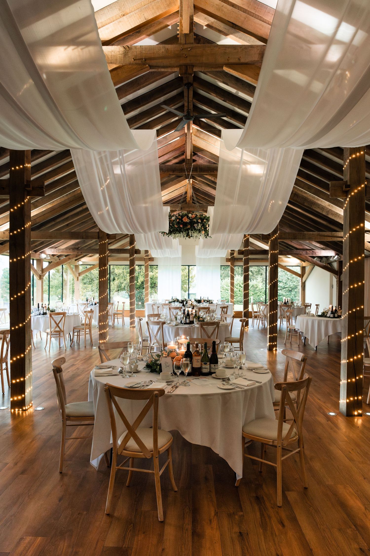 Elegant wedding reception venue with exposed wooden beams, white draping, string lights and rustic cross-back chairs.