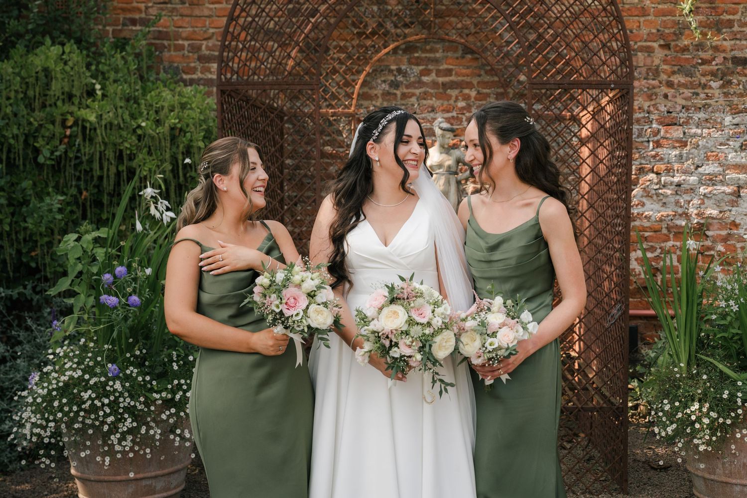 Three women in green bridesmaid dresses and white wedding gown pose together in front of rustic brick archway with flowers.