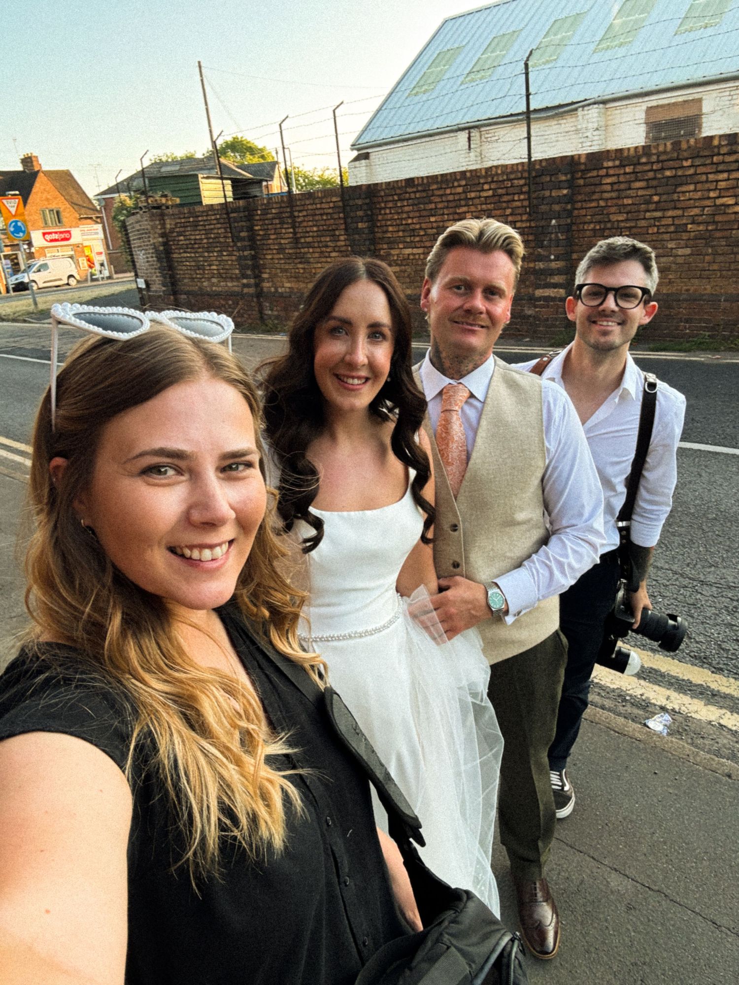 A group of four people posing for a selfie outdoors, including a bride in white dress with wedding party on a street.