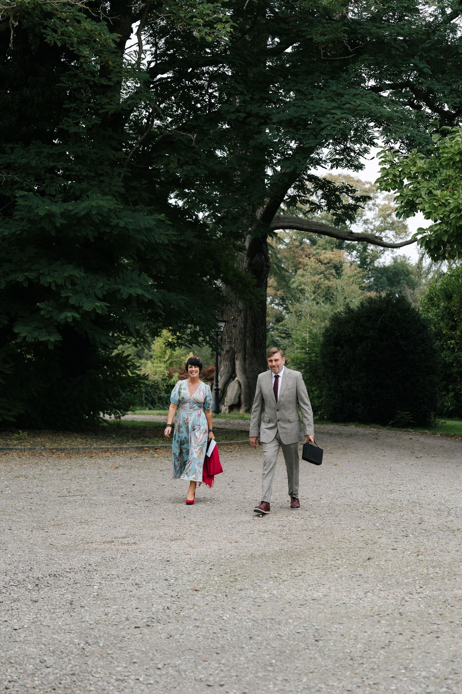 Three people walking on a gravel path through a wooded park area on a sunny day.