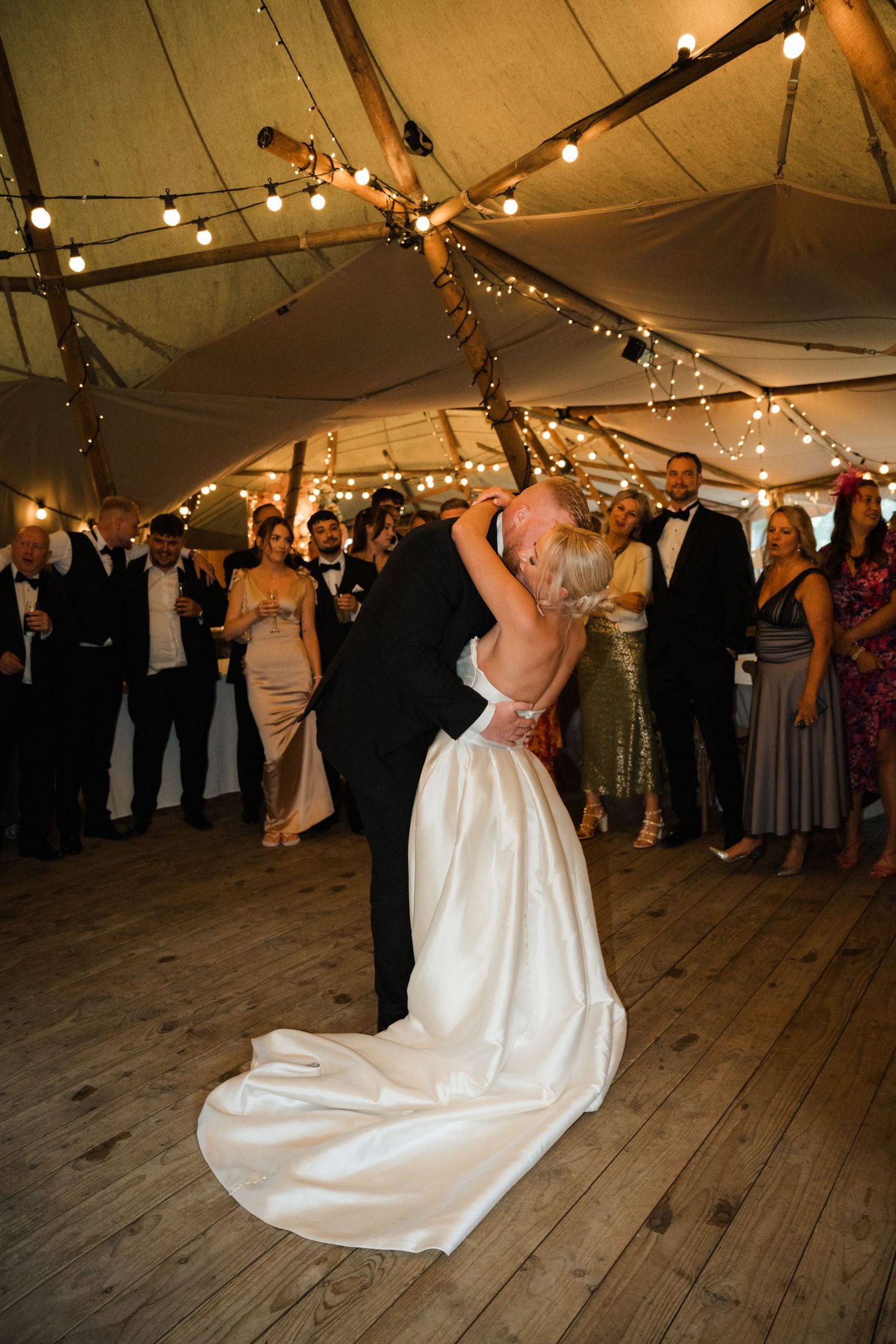 A romantic dance under string lights in a rustic tent venue with wooden floors and gathered wedding guests.