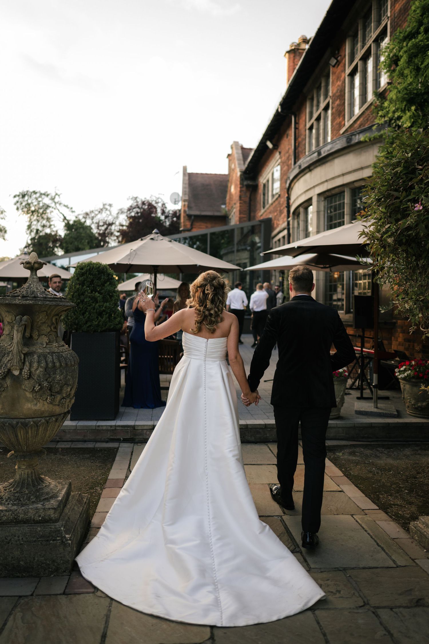 Wedding party walks along stone terrace with brick building and patio umbrellas in background.