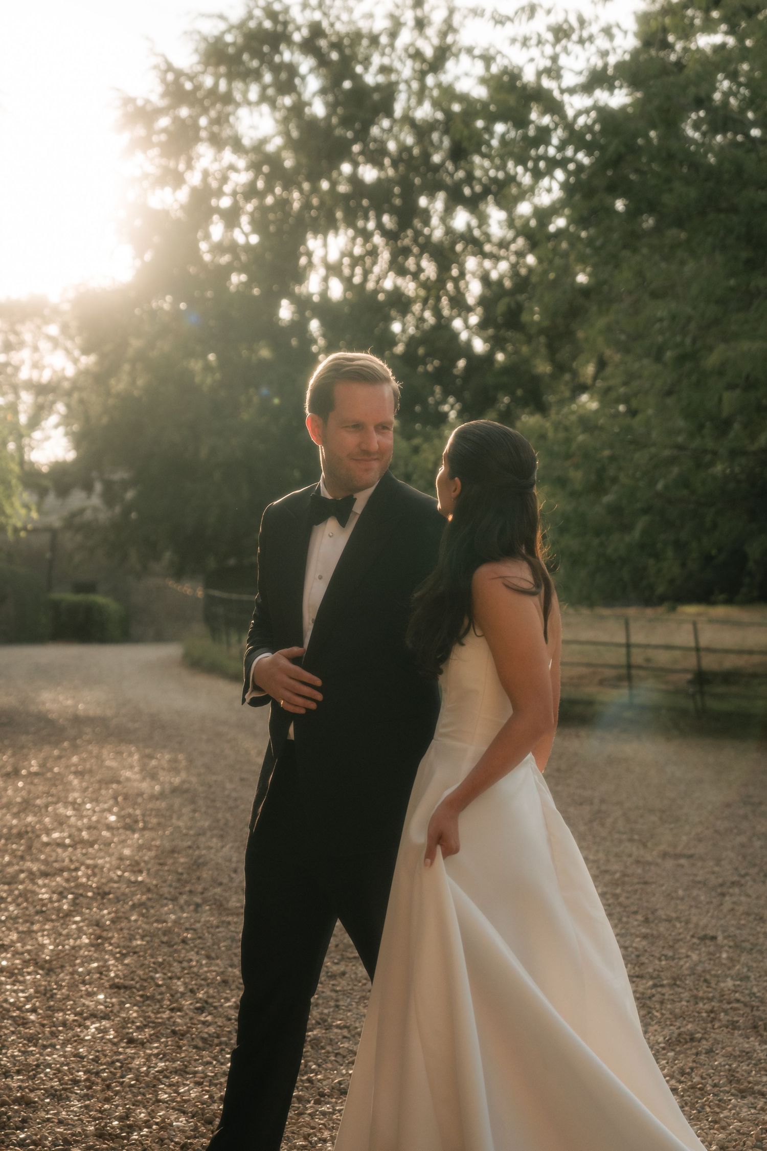 Bride and groom share a romantic golden hour moment on a gravel path, she in a white gown, he in a black tuxedo.