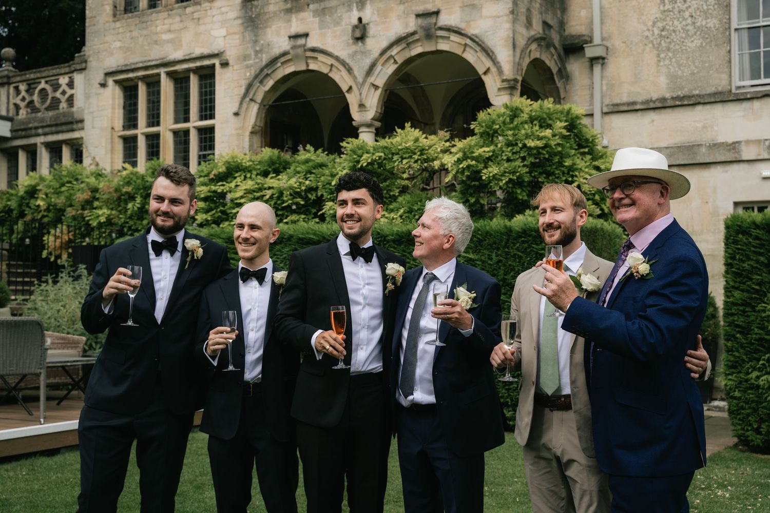A group of formally dressed guests enjoying drinks together on the lawn of an elegant stone building with arched architecture.