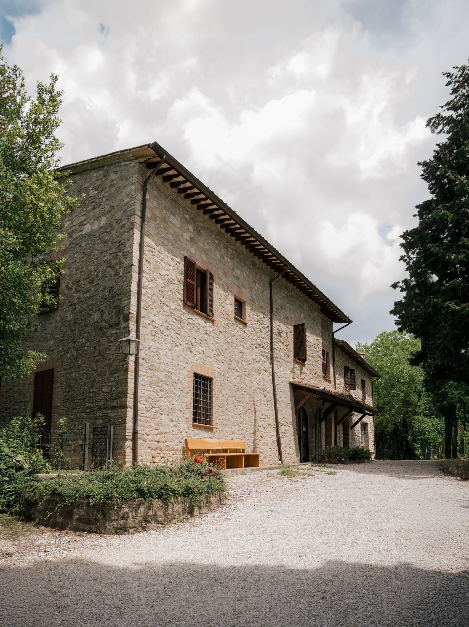 A rustic stone farmhouse with wooden accents and gravel driveway nestled among trees under cloudy skies.