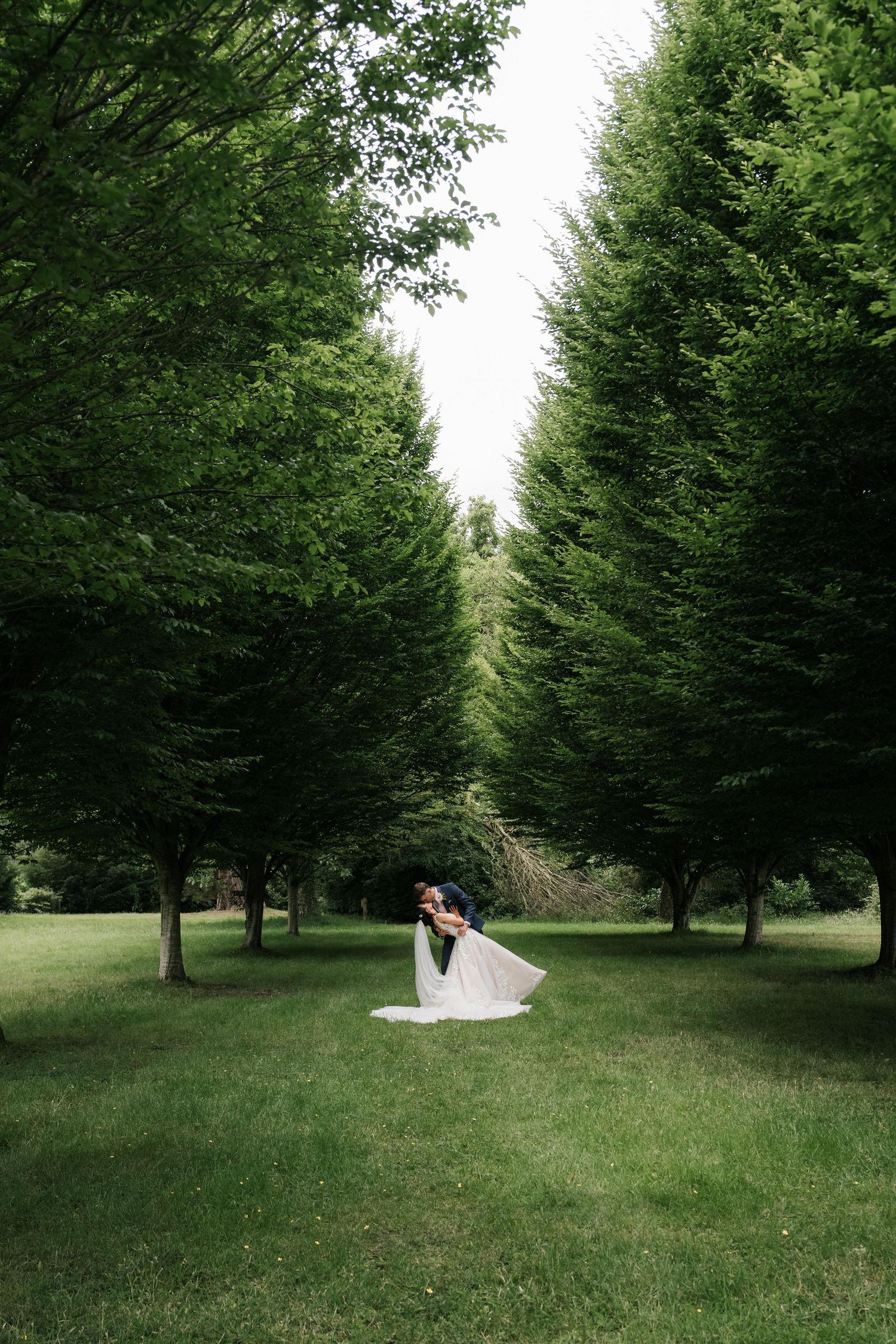 A couple embraces in a romantic moment surrounded by tall evergreen trees in a lush garden setting.