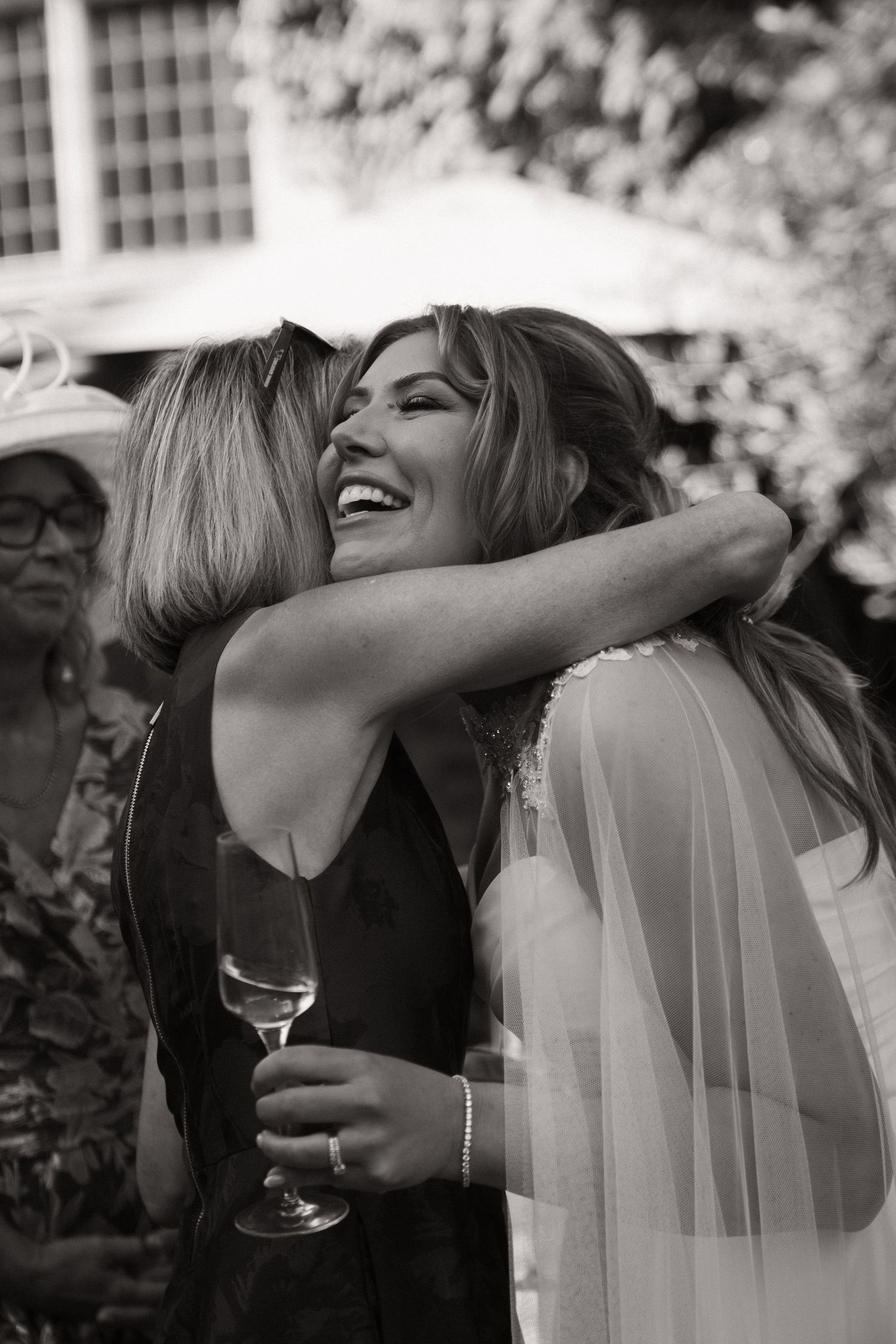 A black and white candid photo shows a joyful embrace between two people at a wedding celebration, with a veil visible.