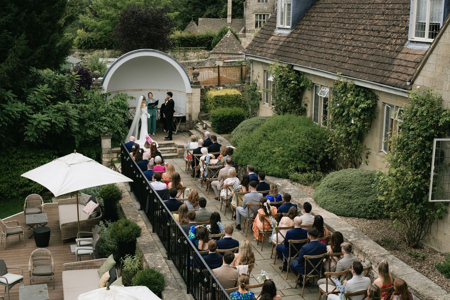 Guests seated outdoors at a garden wedding ceremony with stone house and greenery in the background.