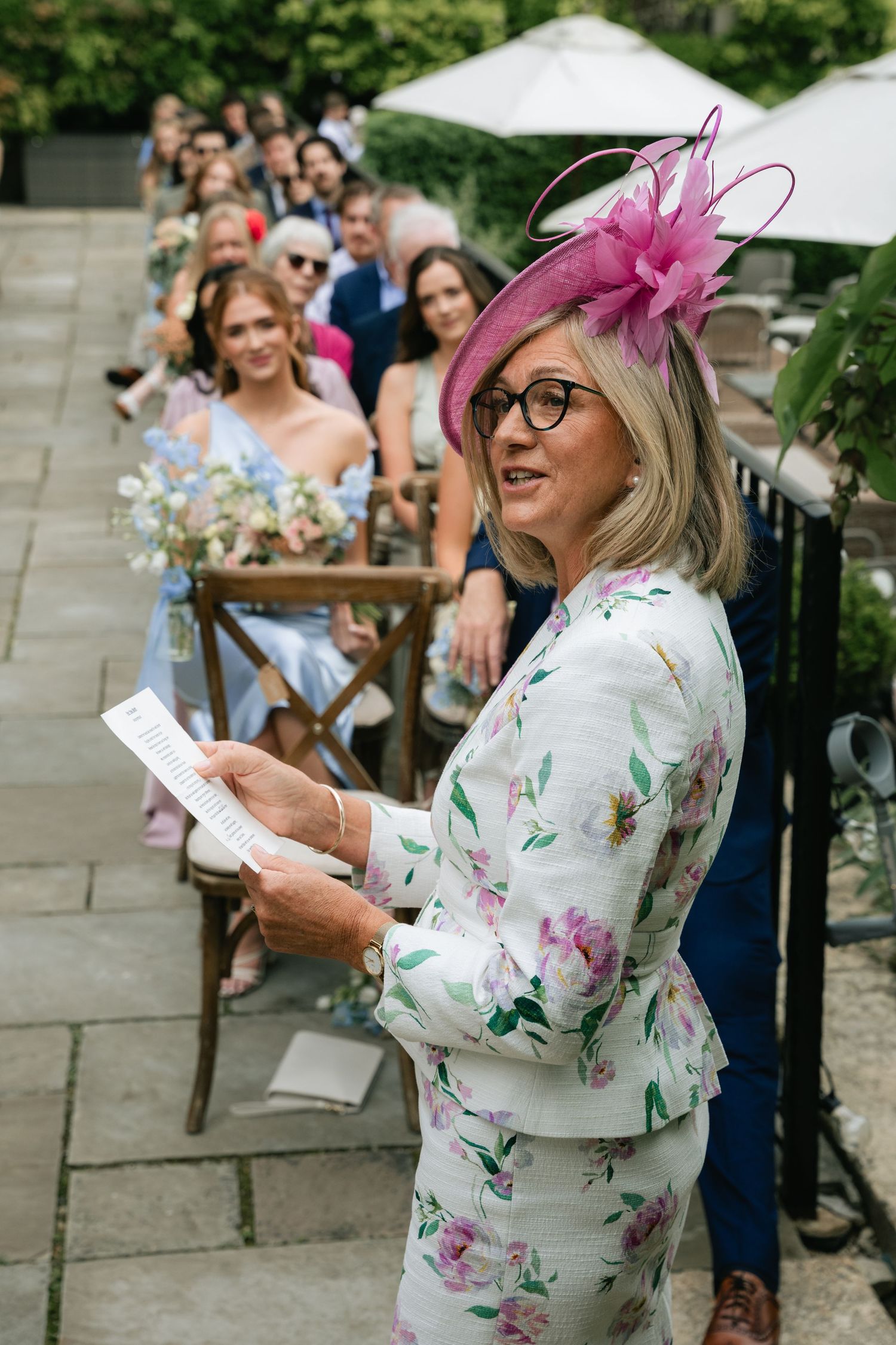 A guest in a floral dress and pink fascinator reads from a paper at an outdoor wedding celebration.