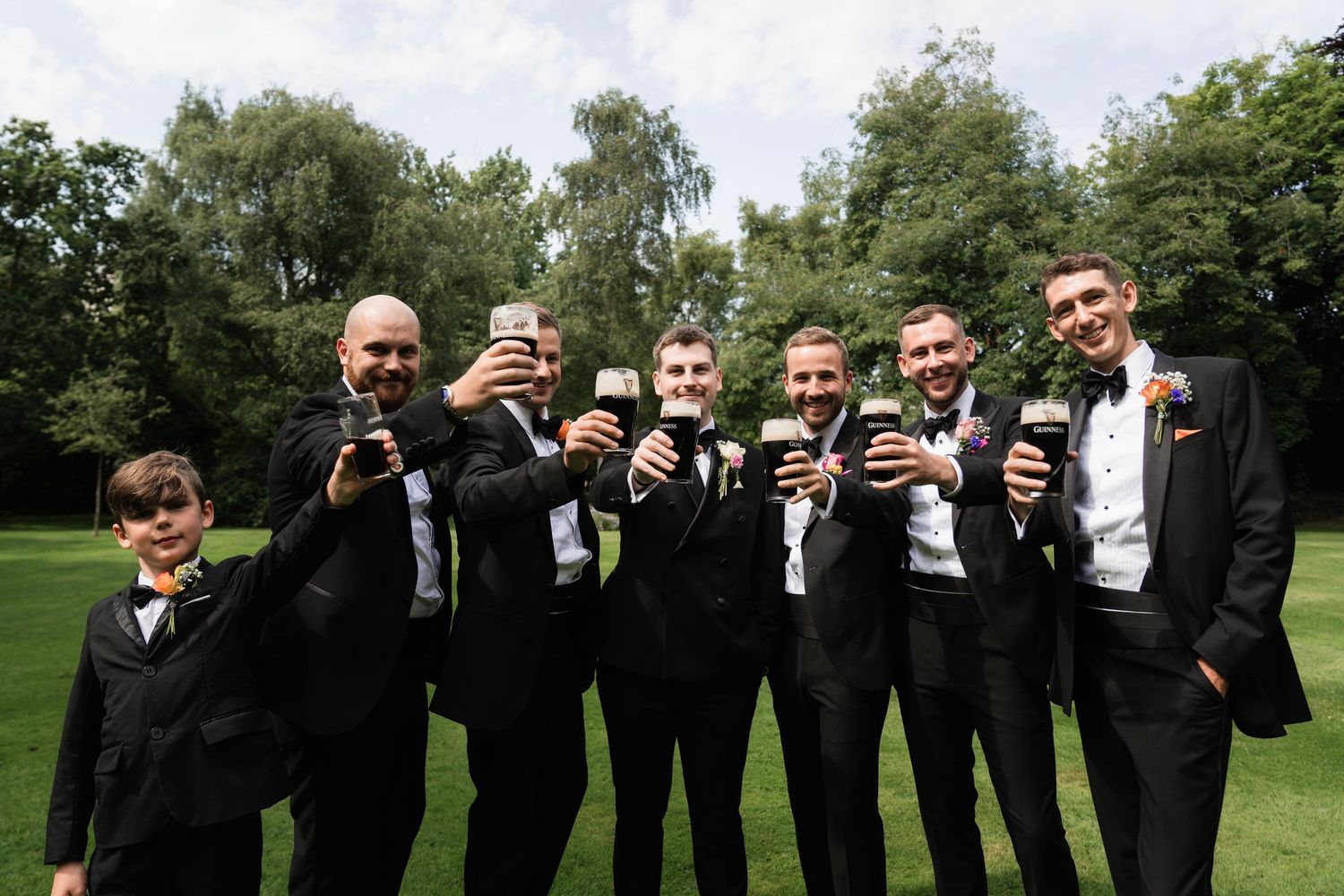 Wedding party groomsmen in black suits strike fun poses while holding drinks outdoors on a golf course.