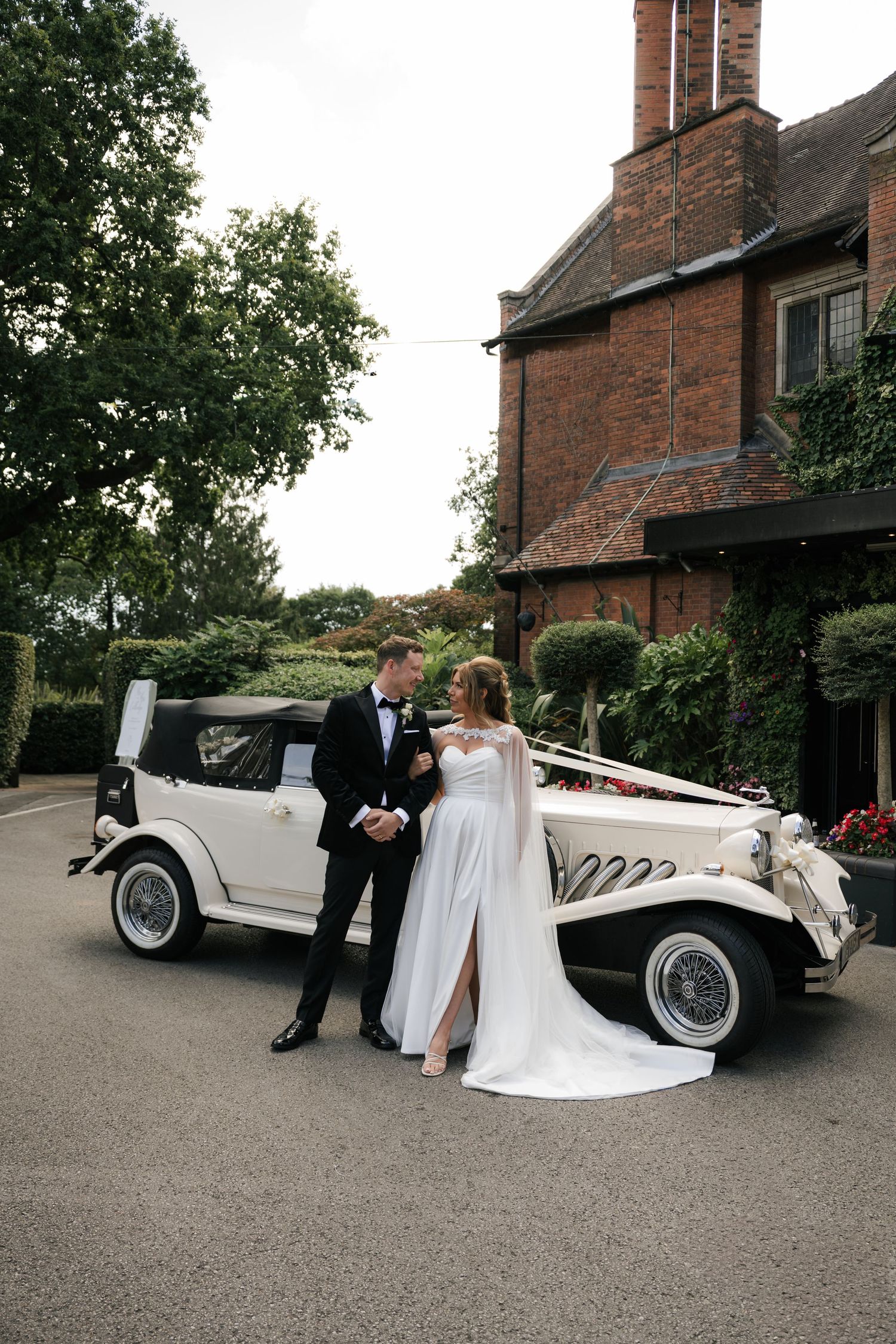 Classic white vintage car parked outside brick house with wedding couple posing beside it on driveway.