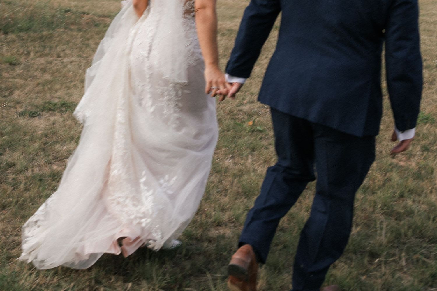 A bride in a flowing white lace gown and groom in navy suit walk together on dry grass in a rustic outdoor setting.