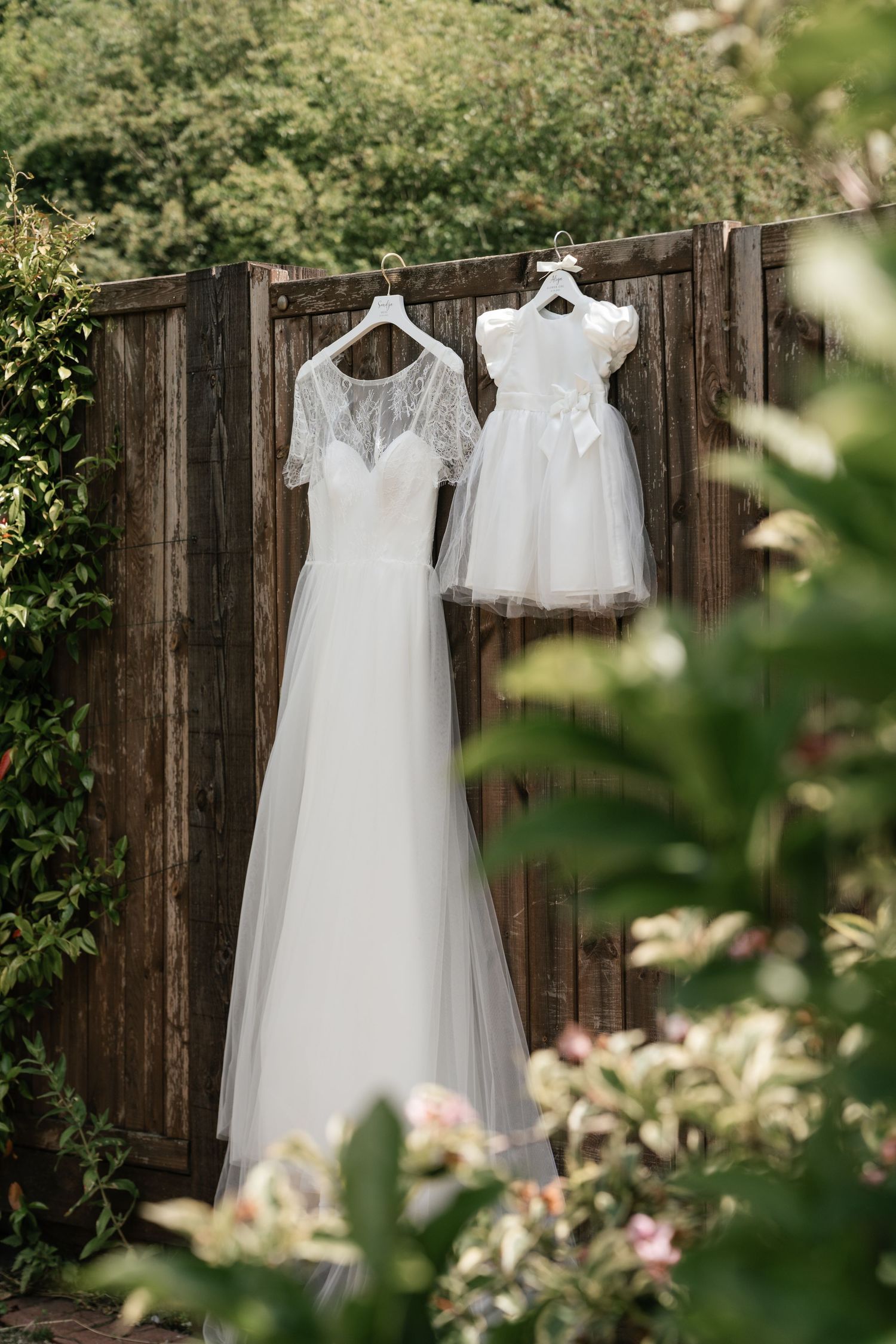 White wedding dresses hanging on wooden hangers against a rustic fence with greenery in the background.