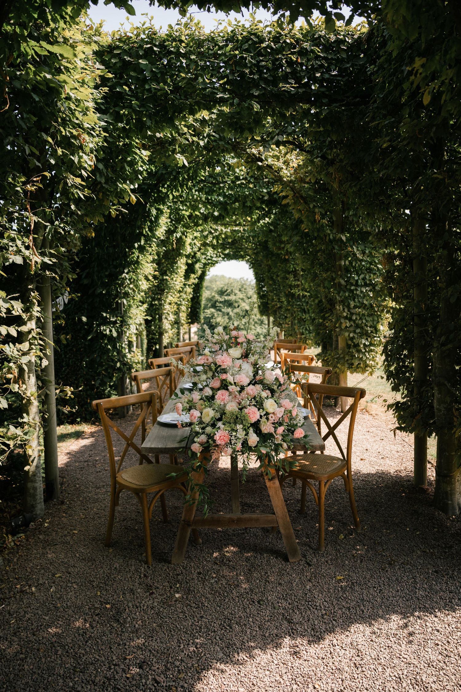 A rustic outdoor dining table with wooden chairs set under a lush green garden archway with pink floral centerpieces.