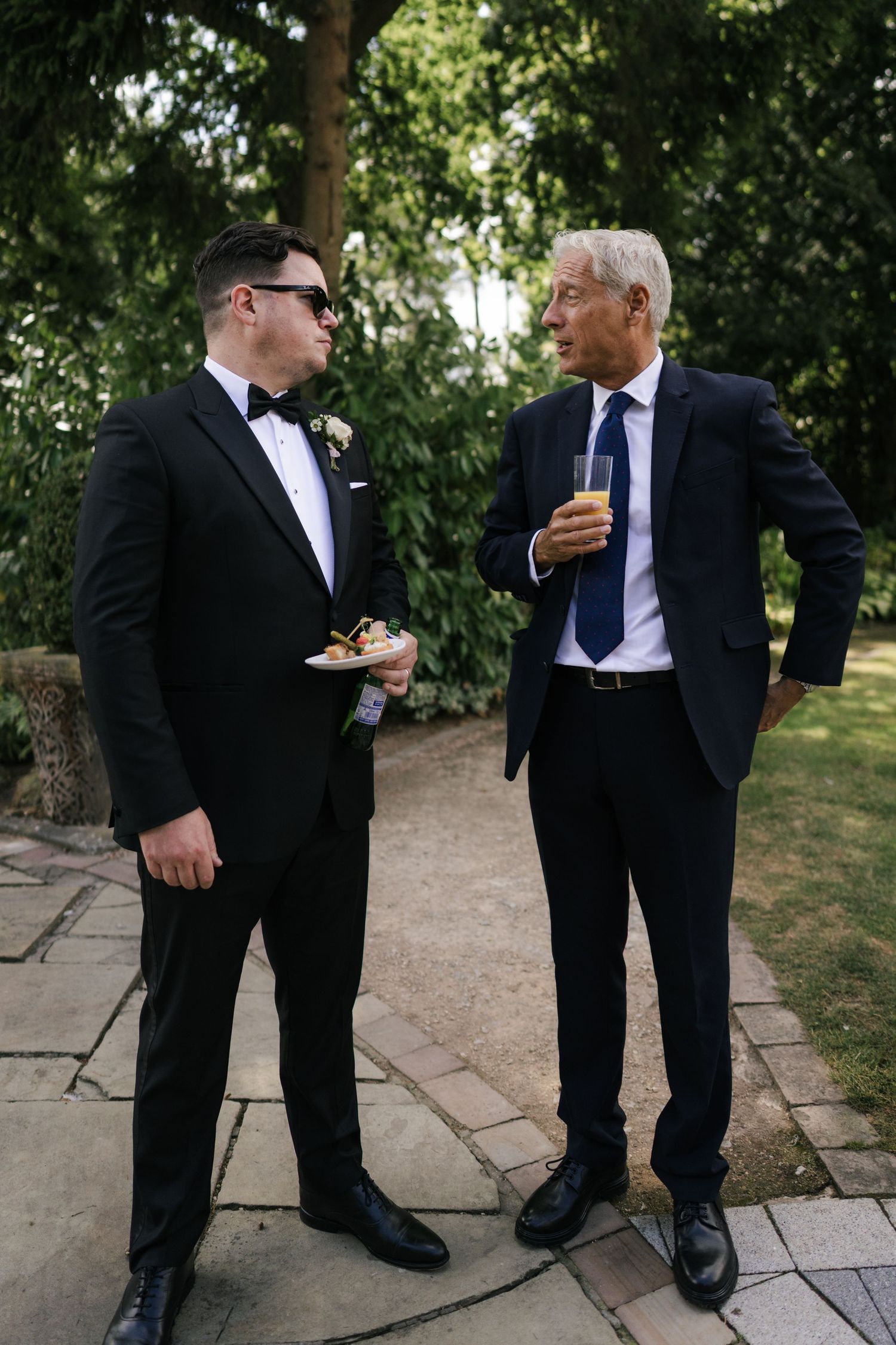 Two men in formal attire converse while standing on a stone path in a garden setting.