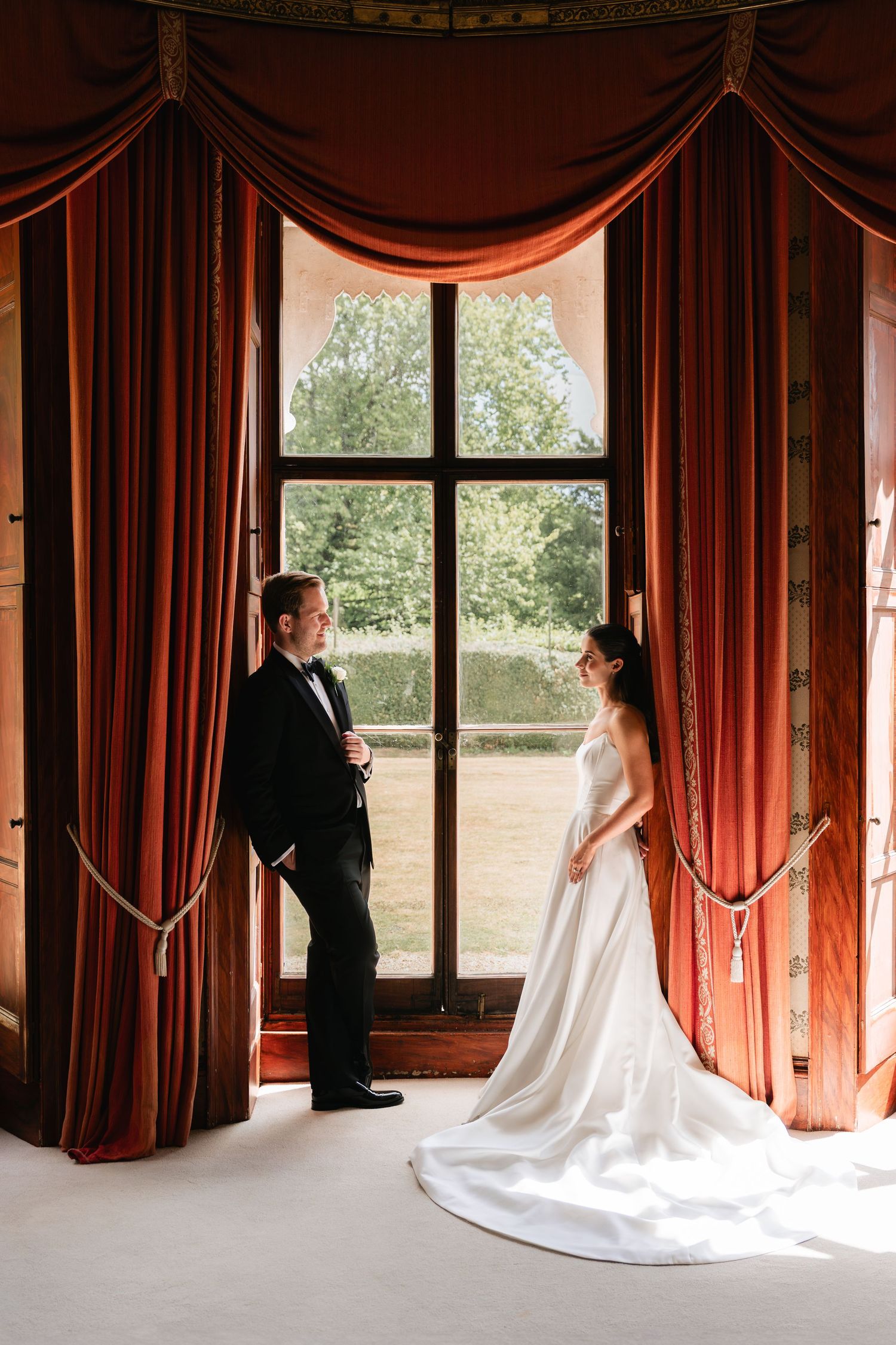 Bride and groom stand facing each other by a grand window with red velvet drapes in an elegant wedding venue.