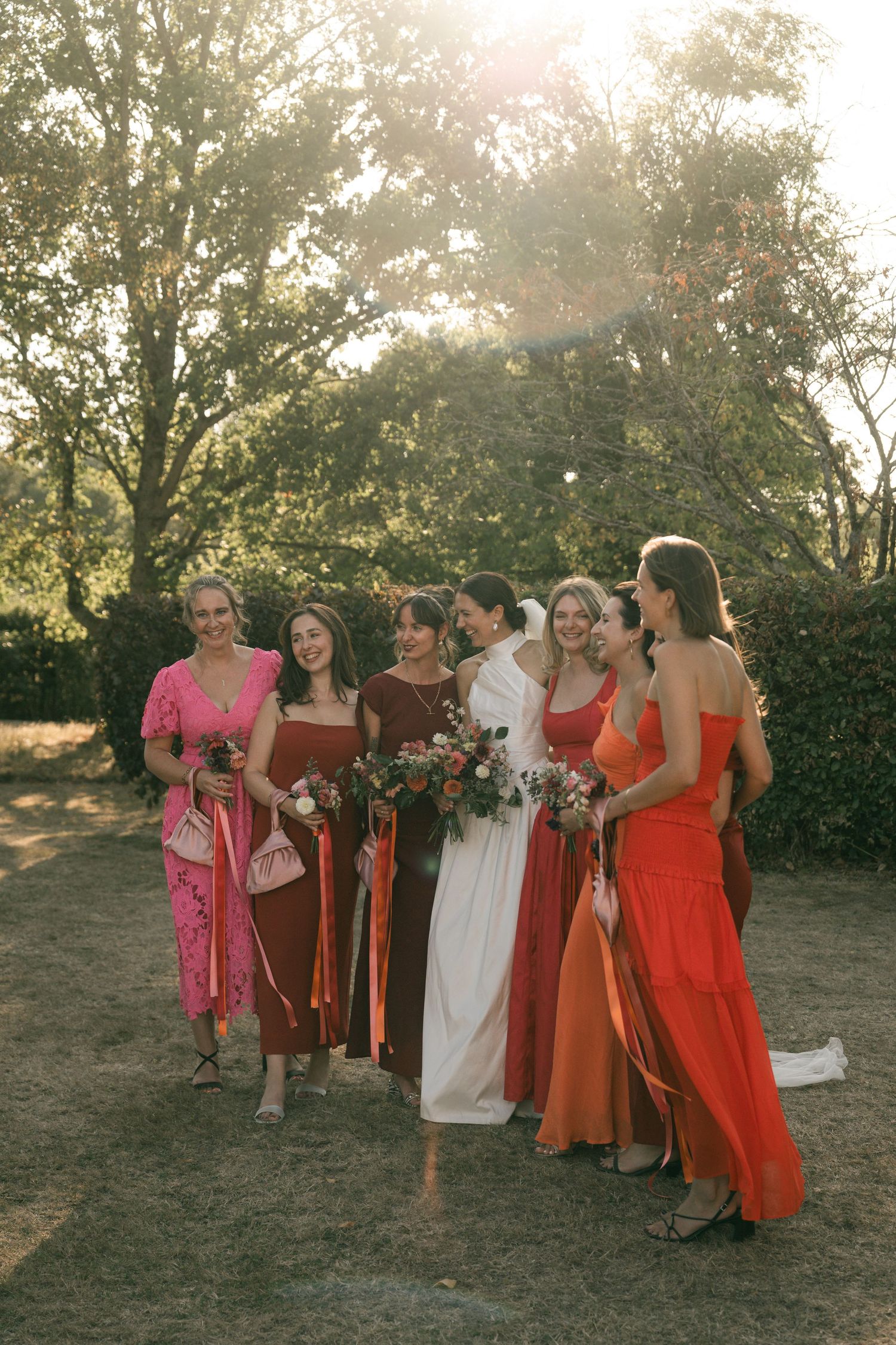 A group in orange and pink dresses poses together outdoors at sunset during a wedding celebration.