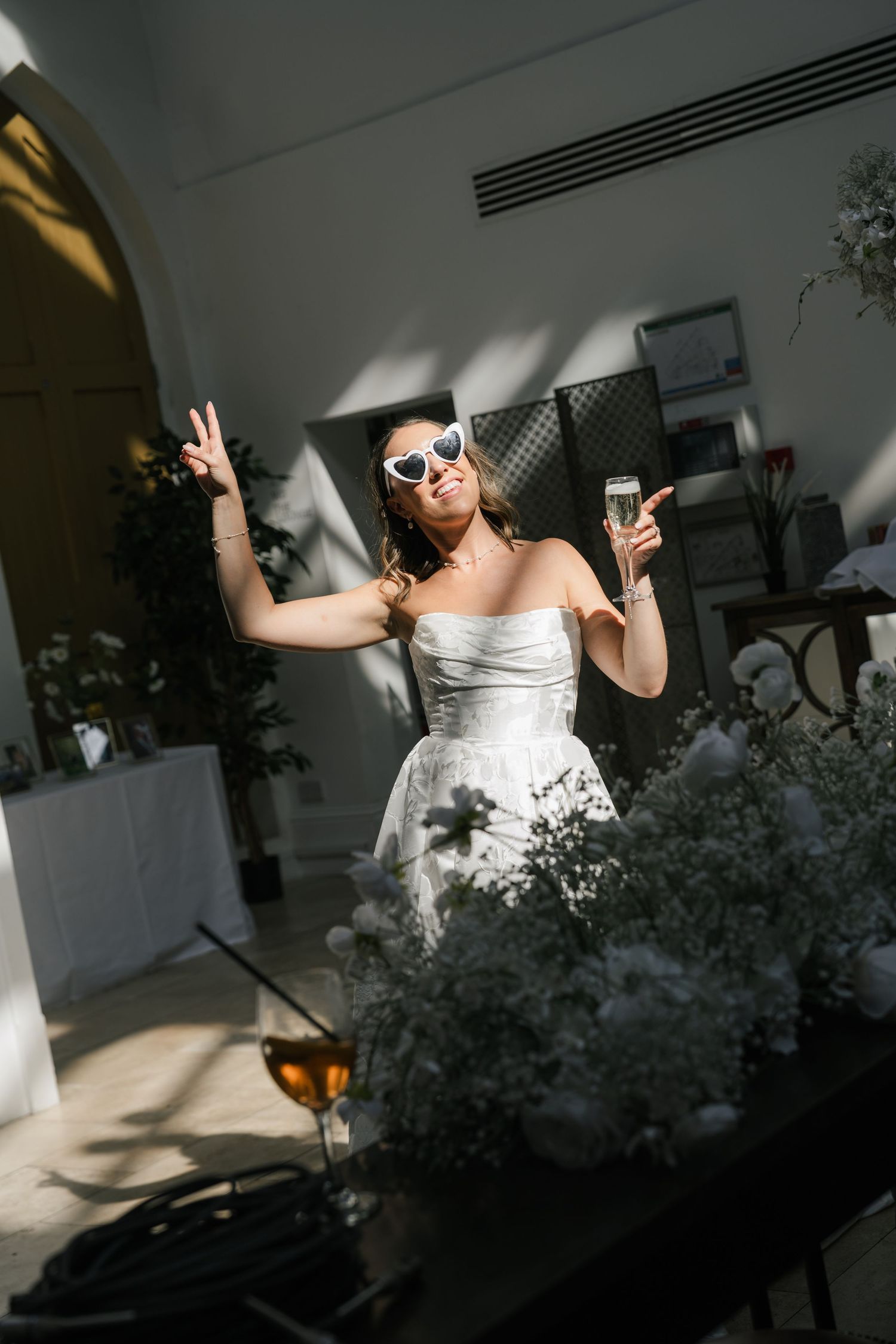 A joyful person in a white wedding dress and sunglasses dances while holding a champagne glass in sunlit room with greenery.