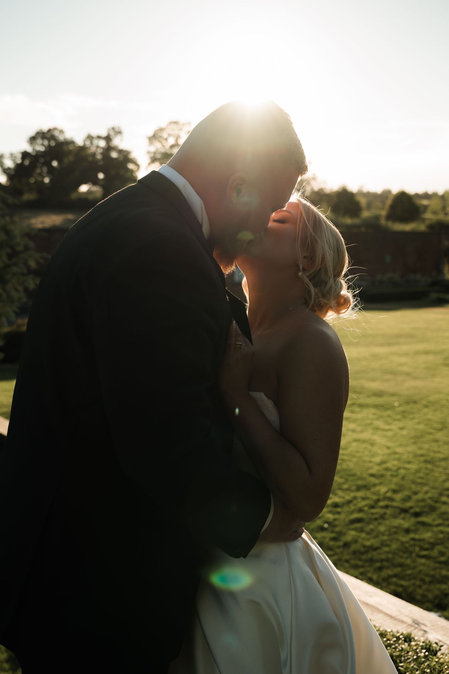 Silhouetted couple shares romantic kiss at sunset in grassy field with golden sunlight glowing behind them.