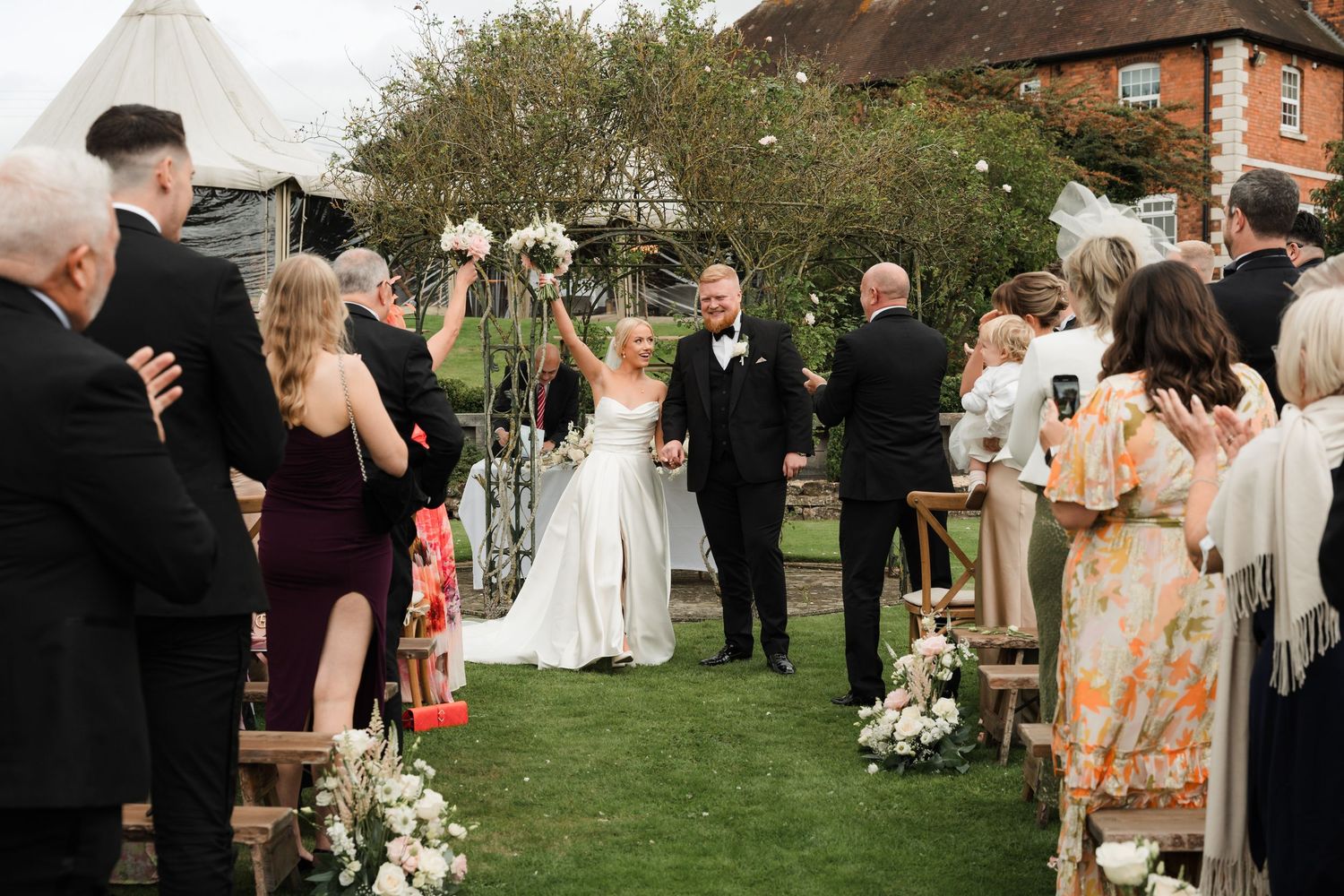 Wedding guests gather outdoors on a lawn near a rustic brick building during an elegant countryside celebration.