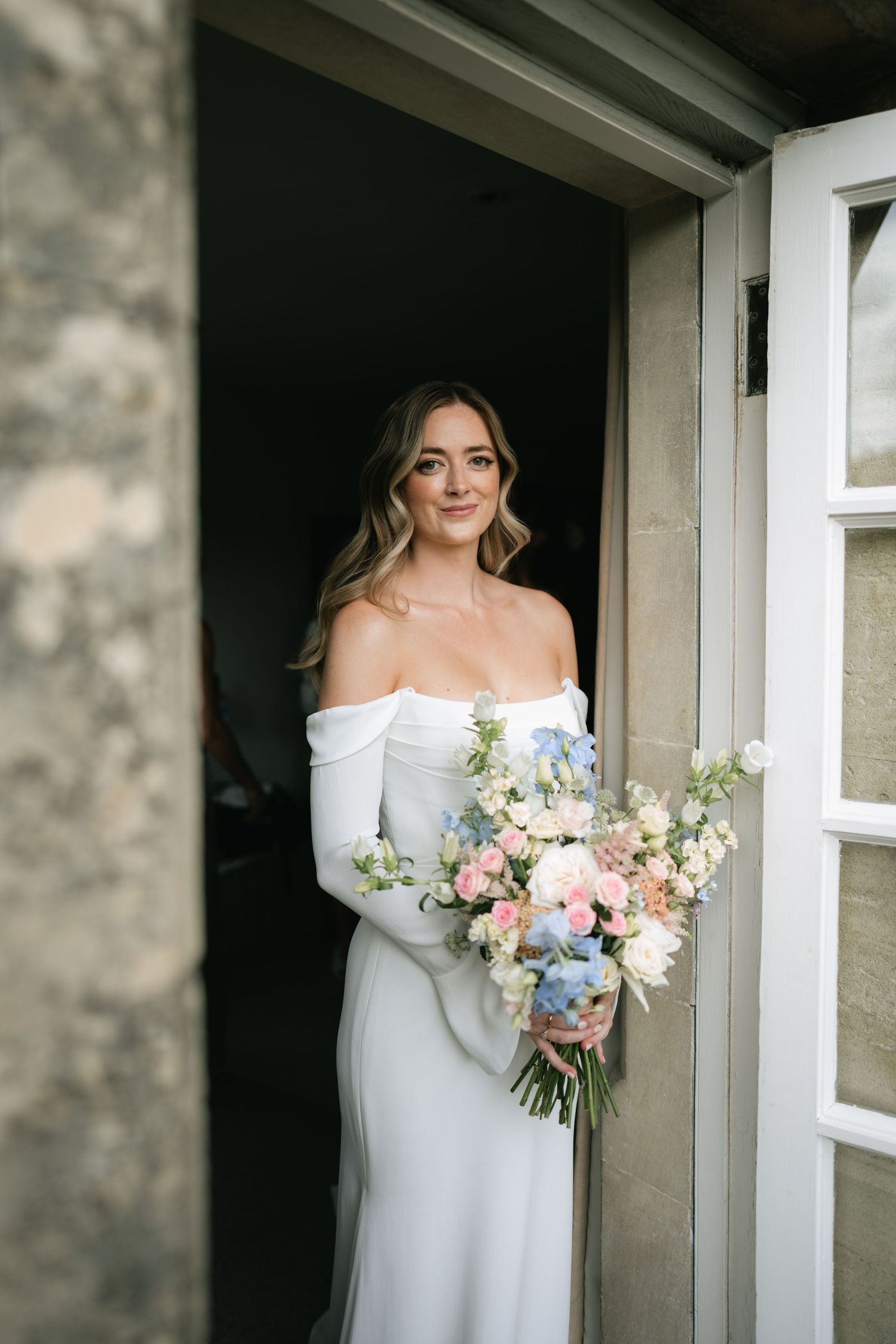 Bride in elegant off-shoulder white gown holds delicate bouquet of pink and white flowers by doorway.