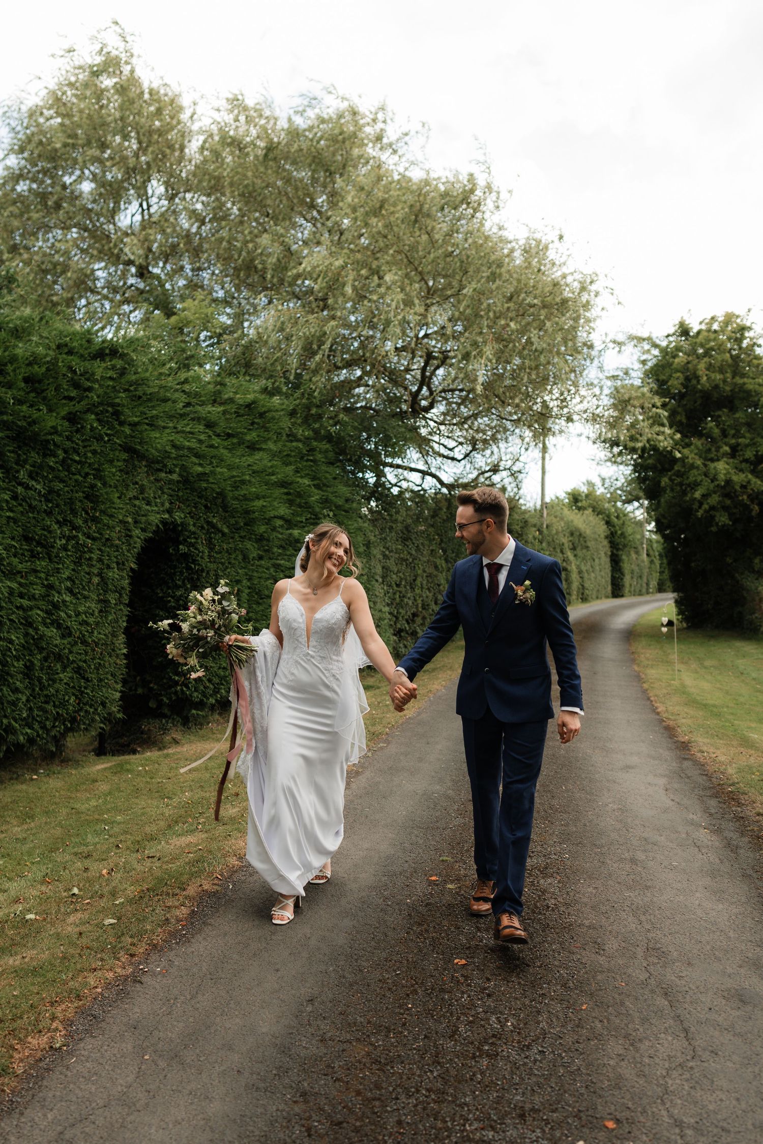 A couple walks together on a gravel path surrounded by green trees and hedges on a wedding day.
