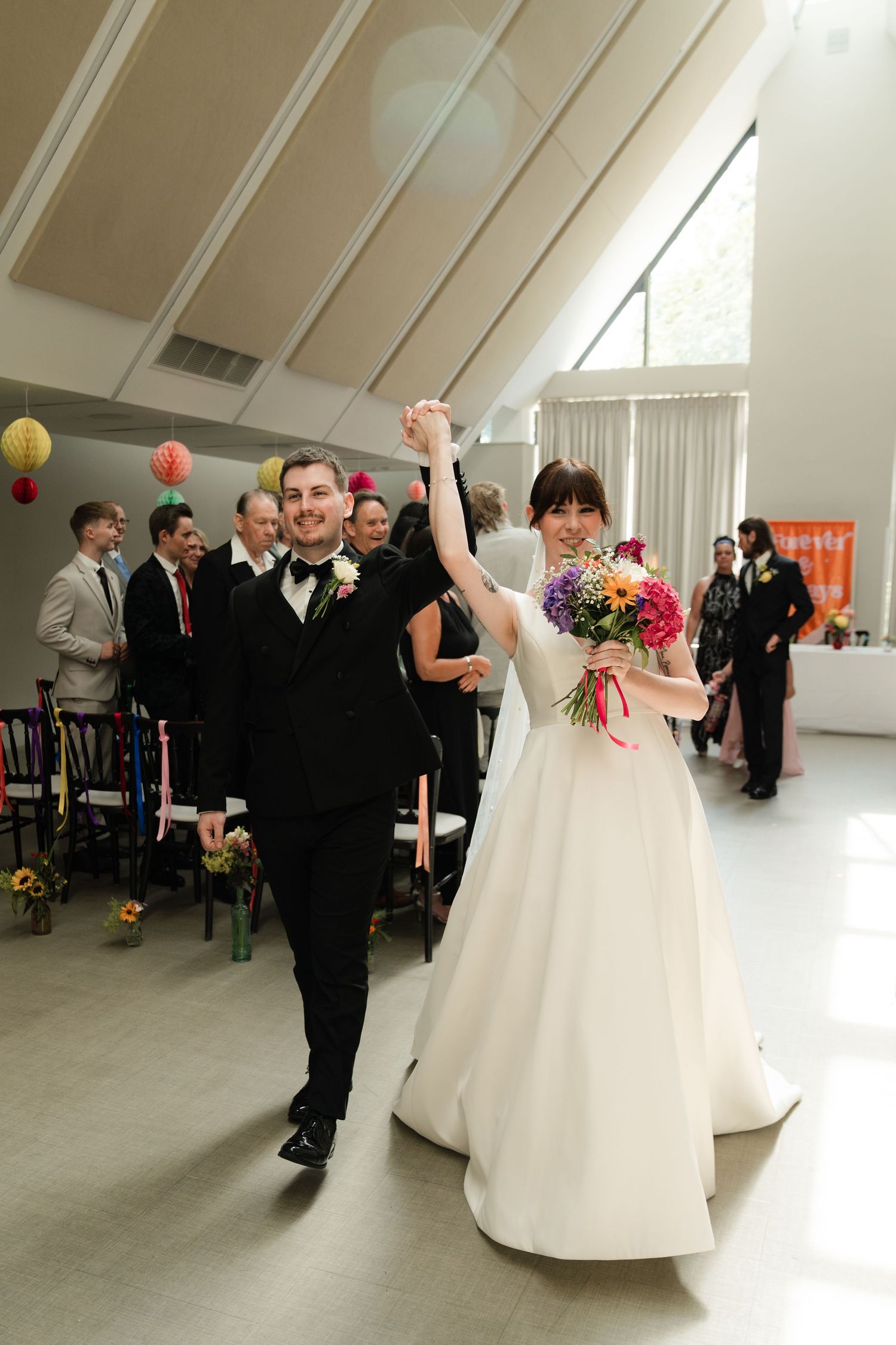 Wedding reception guests gather in a bright white venue with colorful paper lanterns and floral decorations.