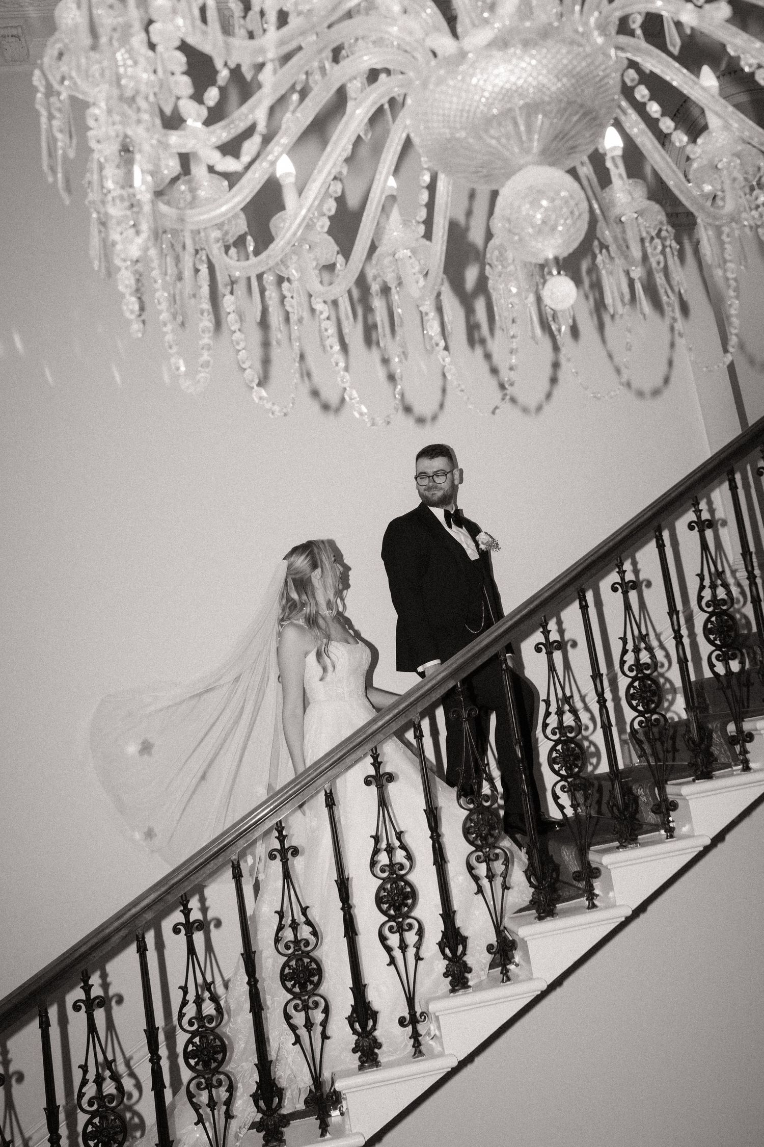 A couple stands on an elegant staircase under a crystal chandelier in a black and white wedding photograph.