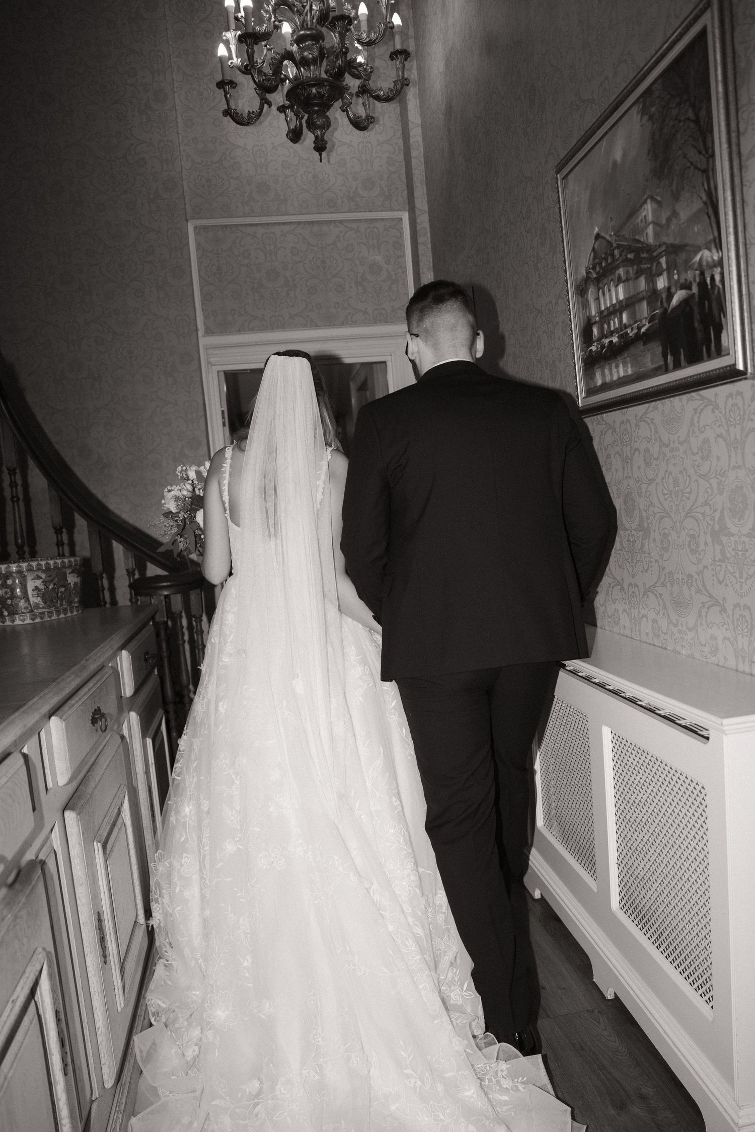 Black and white wedding photos show bride in flowing white gown with long veil walking up stairs with groom in dark suit.