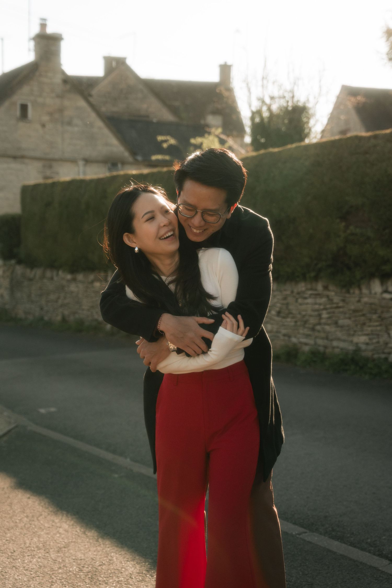Playful couple embracing on street with stone buildings and red trousers visible.