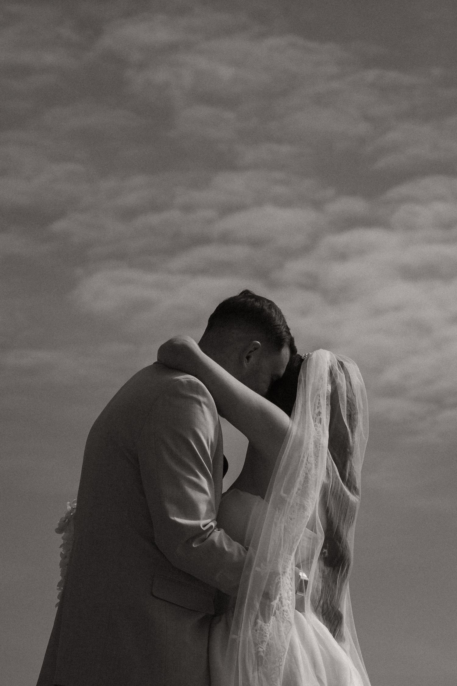 Black and white photo of a bride and groom sharing a romantic embrace under a cloudy sky on their wedding day.