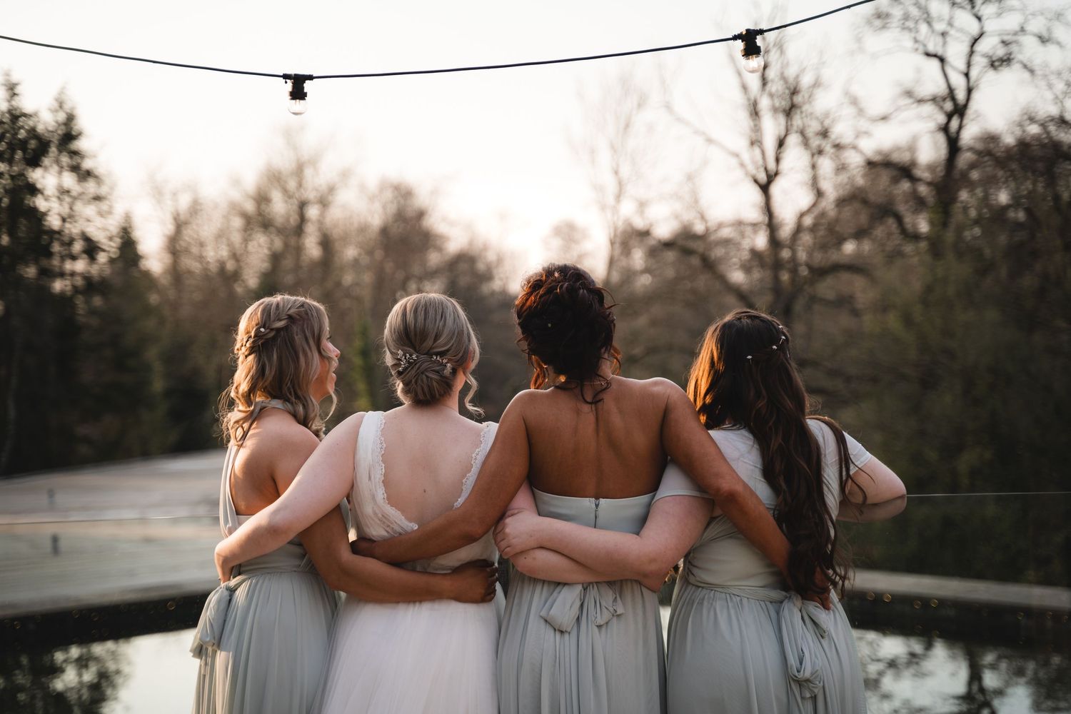 A group of bridesmaids in light gray dresses embrace while looking at a sunset view by a lake.