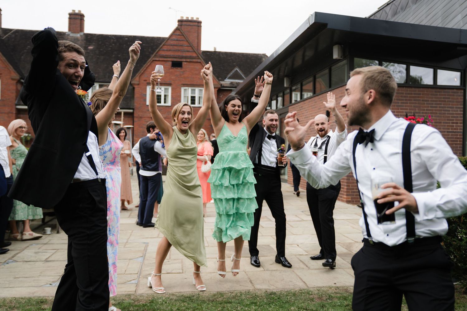 Wedding guests dancing and celebrating outdoors in front of a brick building during a festive reception.