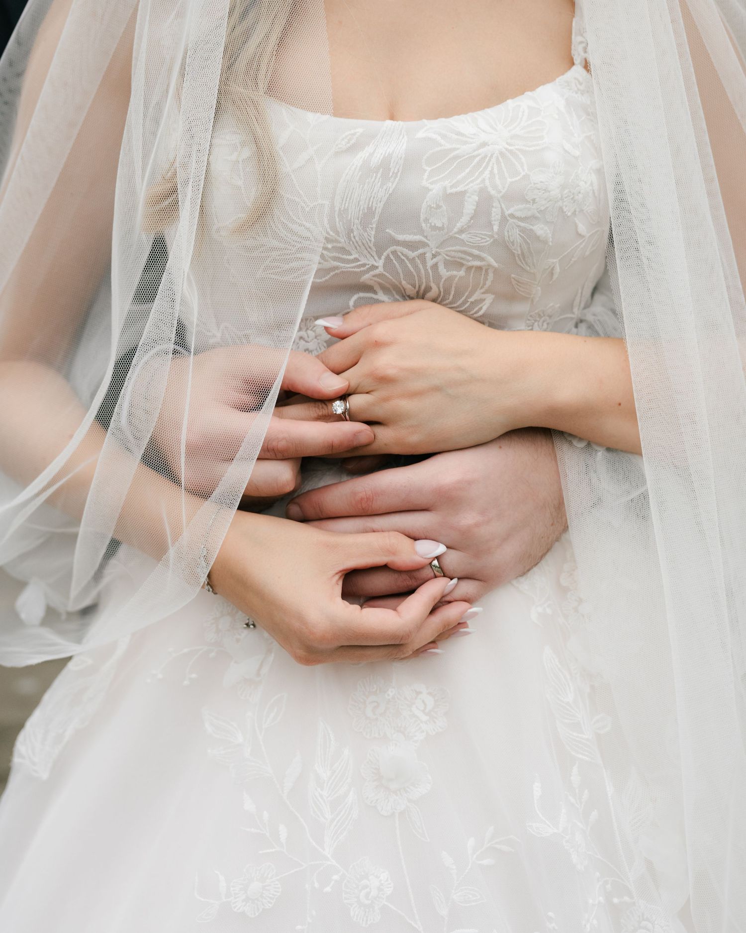 Close-up view of hands resting on a white wedding dress with delicate lace details and tulle veil.